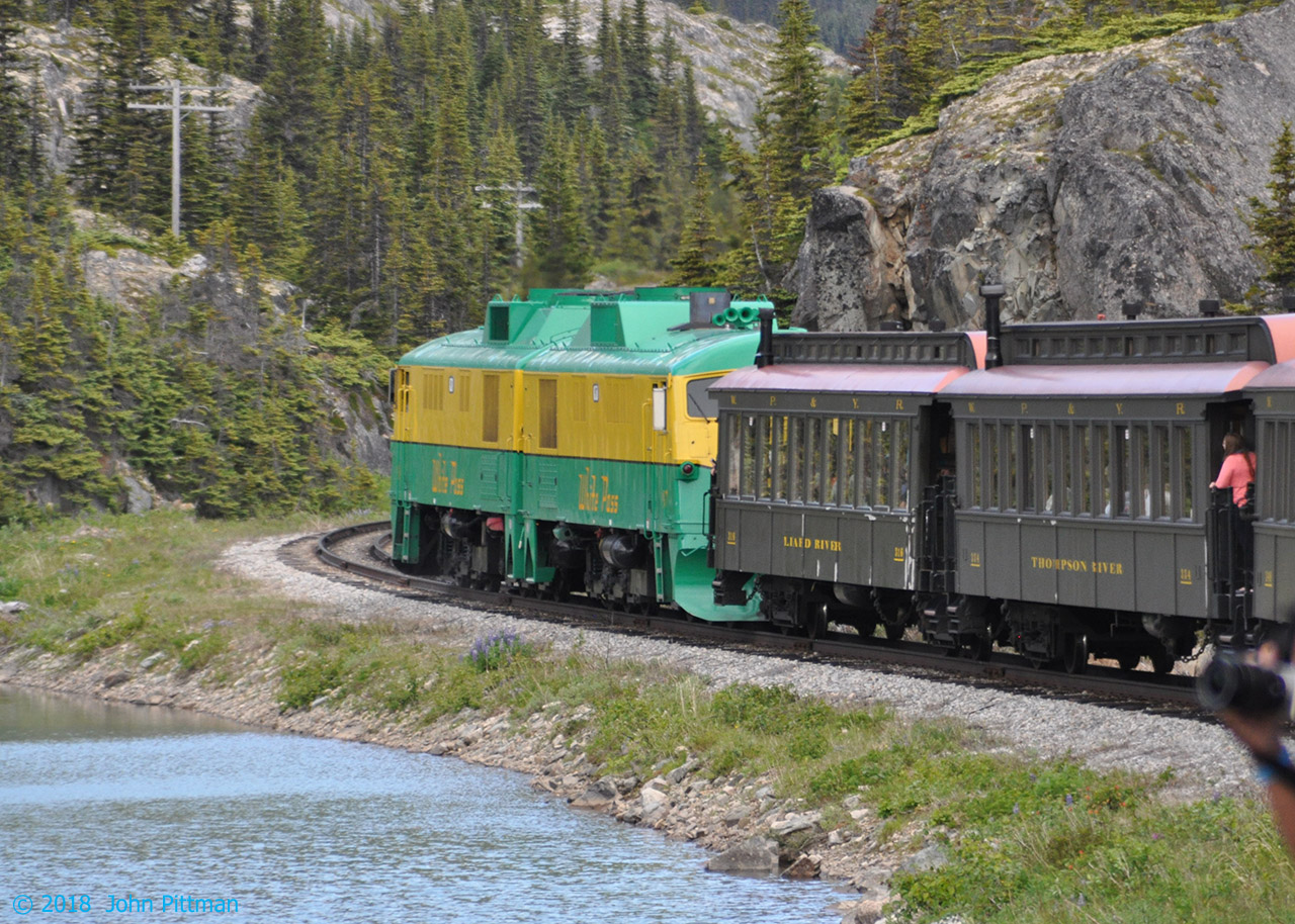 Immediately behind WP&YR GE locomotives 91 and 97 (GEX3341 had Alco 6-251B, repowered Cummins QSK45L) are coaches 316 Liard River, and 334 Thompson River. Now over the 873m White Pass Summit and into BC, the railway descends more gradually toward the level of Bernard Lake near Fraser BC.  In between mountains, the terrain is heavily glaciated (gouged) with parallel lakes and areas of thin soil and bare bedrock.