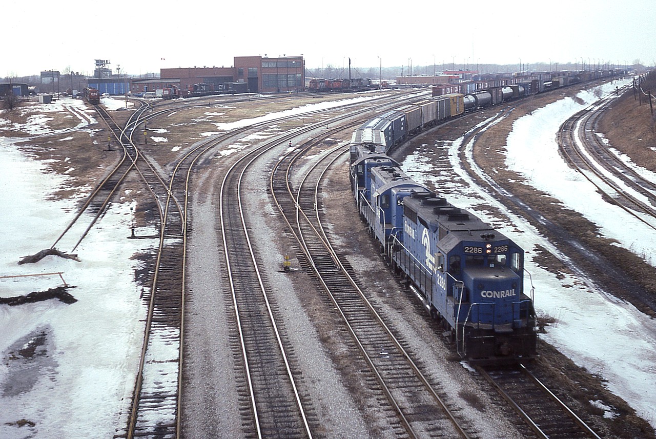 (Photo 1 of 2) This is the sprawling CN facility at Fort Erie as it looked in early 1984. One can see the turntable and roundhouse in extreme left background, toward the centre the CN shop building; capable of extensive repairs, and to the right of that, fueling area, CN offices where paperwork and scale records kept; and on this day very little traffic in the yard. Lower right is the mainline. CR 2286, 5435 and 7548 have a fair sized train to return via the International Bridge, after bringing in Canadian traffic a few hours earlier. Photo taken from Central Ave bridge.
Yeah, it is all gone now but the mainline. The CN main shop building still stands, rather forlorn looking, as the home of the fledgling Niagara Railroad Museum. It is worth a visit.