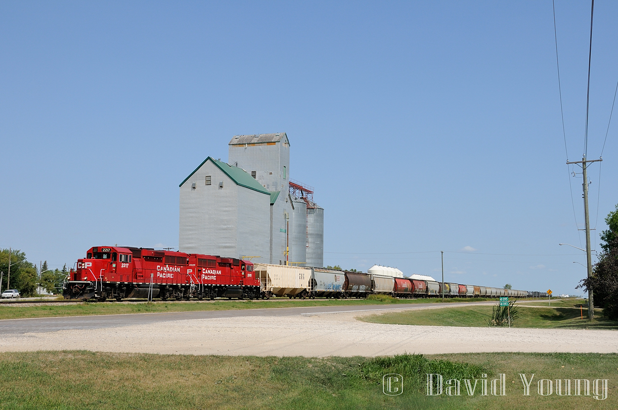 Westbound (in actuality southbound) P03-20, "The Altona" rolls through the small southern Manitoba town of Domain, located along Provincial Road 330. With 47 cars in tow, none of which are for the local elevator, GP20C-ECO's 2217 and 2325 have their train in hand as they hustle along at track speed.