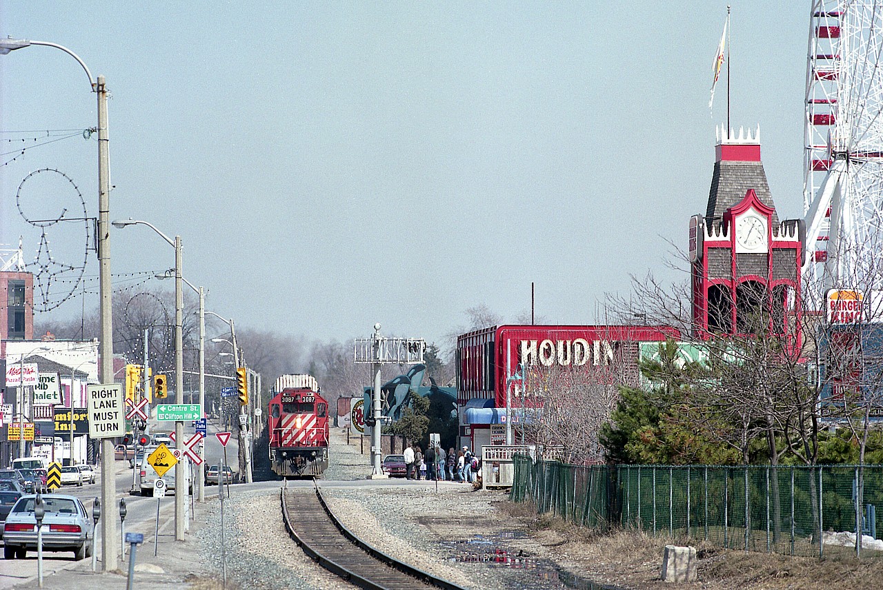 A trio of CP GP38-2 locomotives pull traffic from the USA en route to Toronto. Units are 3087, 3082 and 3062. Train is just coming up to Clifton Hill tourist mecca, where it will snarl traffic as the slow moving train makes its way thru the city. In the background one can see, curving from the left, the autoracks as the train parallels Palmer Ave back there along Mile 1. This image is interesting as it shows so much of the area at that time. Billboards on the right behind Houdini's include Marineland and Yuk-Yuks. The tower clock reads 1235, about right, so perhaps the clock actually works! One can see also on the right where the former second track used to be. Victoria Av on the left used to be part of Ontario Kings Hwy 20, but changes by the Mike Harris government, shedding themselves of responsibility, left the maintenance to the city. The Houdini structure actually used to be a railroad station many years ago......whose? Ferris Wheel on the right is gone, along with all the rest of Maple Leaf Village. Niagara Falls is ever-changing. Its a jungle out there...............and no more railroad. It lasted another 8 years or so.