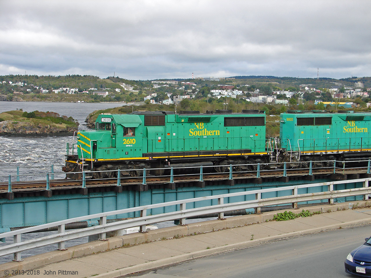 New Brunswick Southern GP38-3 units 2610 and 9802 lead a freight train west over the ex-CP bridge that crosses the lower St John River. The engines are just beyond the girder truss section. Vantage point is the St John NB information centre building near the Reversing Falls viewpoint. 
NBSR 2610 was built by EMD in 1965 as GP35 SP 7768. It was upgraded into a "GP38-3", losing its turbocharger and middle radiator fan, presumably converted from 567 to 645 diesel engine and electrically upgraded. It became SP 6665, SP 6308, and CIC 110 (Cedar Rapids & Iowas City). It joined the NBSR roster in 2007, arriving in the UP paint scheme.