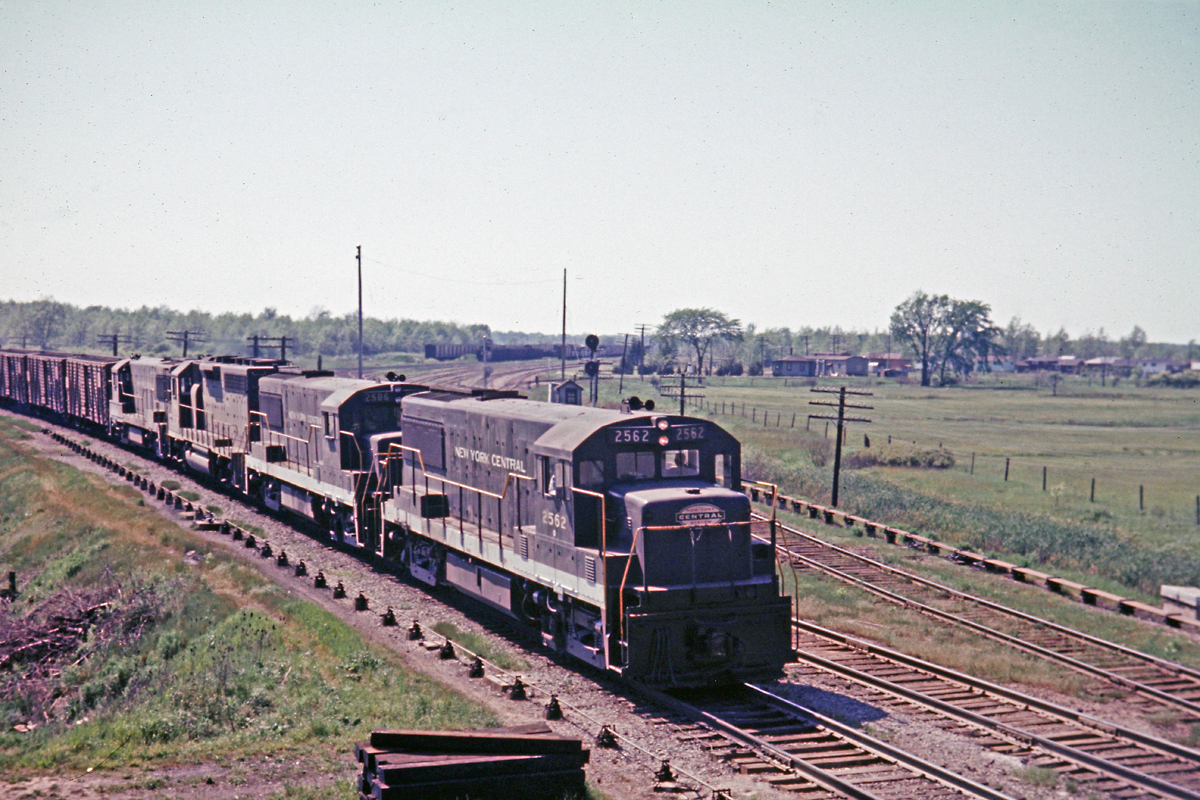 Caption: NY4 with all 4-axle power is seen here on the eastbound approach to Bridge 15 on the Welland Canal in 1967. In the background is Coyle Yard on the TH&B. Notice the interlocking rods on both sides of the tracks. The two tracks of the CASO Sub (MCRR) curve around to the left while the single track on the right is the interchange to the TH&B. Coyle Yard and the CASO Sub are long gone. Bridge 15 still exists and this section of the old Welland Canal is used for recreational purposes now.