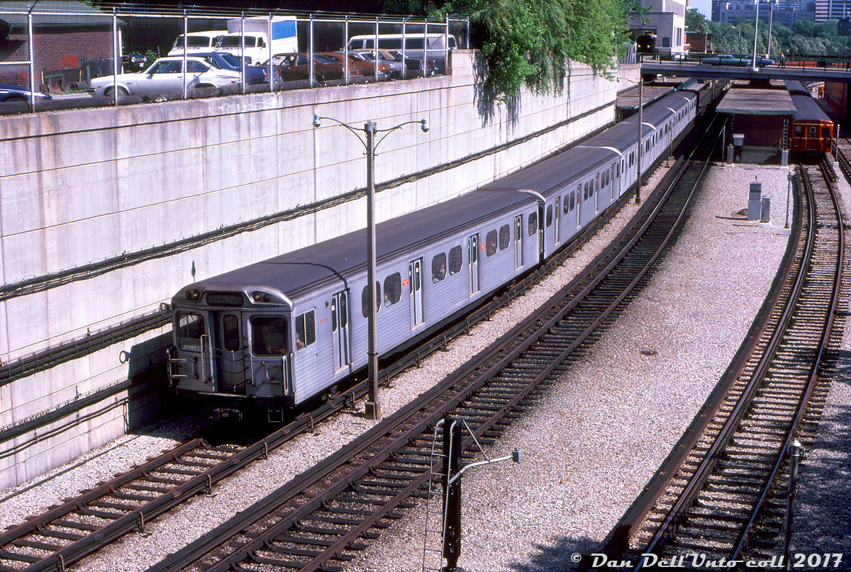 TTC H4 5594 (relatively new at the time, built by Hawker Siddeley Canada in 1974/75) leads a mixed train of H4's and four older H-series cars northbound out of Davisville Subway Station on the Yonge subway line, bound for Finch. An old Gloucester "G" train is sitting on platform 3, possibly waiting to enter service for PM rush hours, and a TTC bus (likely a GM fishbowl) is visible laying over in the bus bays of Davisville station. The lead car 5594 upon its retirement was converted into work car RT-64, and appears to still be on the roster.  Another interesting thing is the "Identra Coil" placed on the top corner of the lead car (sort of like an extra flag would be on a locomotive). Those were used to activate the Next Train sign boxes mounted on the ceiling at station platforms that would display the correct end destination or routing of an arriving train for passengers on the platform (if the train would be short turning, it would show the last station stop for example). One may still be able to find a few of those old sign boxes in place around the system, although by now long out of use.   J. Bryce Lee photo, Dan Dell'Unto collection slide.