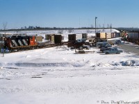 Working a local freight along the Campbellford Sub, CN GP9 4524 is seen switching at the south end of Peterborough, just north of the Highway 7/115 overpass around Ashburnham Drive. Its train sits south of the switch as the lone Geep handles some flatcars at Trent Timber Treating Inc.'s siding around Mile 61. Visible on the right is the local CASE/International Harvester dealer, whose lot is full of various pieces of construction and farm equipment (but who doesn't appear to have been a rail customer at the time). <br><br>CN's Campbellford Sub was cobbled together from various 19th century railway lines and ran from Belleville to Lindsay via Peterborough, but by the time this photo was taken in the mid-1980's, time was running out for many of the unprofitable low-traffic branchlines in the Southern Ontario area including the Campbellford Sub. A year later in 1987, CN would abandon a large section of the line between Corbyville (north of Belleville at Mile 3.2) and the south end of Peterborough (Mile 60.6, a bit futher south of this photo), but continued to operate any local service between Lindsay and Peterborough via the Uxbridge Sub connection in Lindsay. <br><br> Faced with mounting operational losses, CN would pull out of Peterborough for good when it applied to abandon the Lindsay to Peterborough segment, which was approved in <a href=https://www.otc-cta.gc.ca/eng/ruling/298-r-1989><b>June 1989</b></a>. Also included were the abandonment of the Lakefield Spur running north-east out of the city, the Ashburnham Branch on the east side of the Otonabee River (that at one time also served the Quaker Oats plant via a bridge at the north end), and the sale of a number of CN's industrial lines in Peterborough to CP for continued service to the few remaining customers, including Trent Timber Treating (the spurs were then accessed off CP's Havelock Sub). Eventually CP would abandon these former CN industrial spurs in 2012 citing costs and a lack of traffic, and remove most of the trackage around 2015-2016.<br><br><i>Keith Hansen photo, Dan Dell'Unto collection slide.</i>