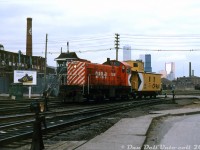 <b><i>Relics of the past:</i></b> CP S2 7026 and van 437227 amble over Strachan Avenue grade crossing, passing the crossing tower as they head north into Parkdale Yard as light power. At the time a gateman manning the crossing tower still manually operated the Strachan Ave. gates and warning lights for the CN Weston Sub and CP Galt Sub that crossed here on their way to the Toronto Terminals Railway territory downtown (the green shack near the Adidas sneaker ad was apparently the gateman's loo). Coloured switch lanterns mounted on switchstands dot the south end of Parkdale Yard's tracks. Visible in the background are the old John B. Smith & Sons building on the left (a former lumber dealer), the city's Wellington Destructor (garbage incinerator) on the right, and two tank cars spotted on the siding for the Toronto Municipal Abbatoir / Quality Packers ("tank siding", off of CN's Weston Sub). Modern office towers in the distance (and CP's Royal York Hotel) contrast with the gritty blue-collar industrial environs of the Parkdale area.<br><br>One of many small switchers built by Alco (and later MLW) for CP, the little 1000hp 7026 dates back to 1945 when CP was still ordering brand new steam engines (for road service) alongside diesel yard switchers. By 1979, the motive power assignment sheets show 7026 was one of nearly 30 little S2, S3 and S4 switchers assigned and maintained out of John St. Roundhouse downtown for local and yard switching duties in the Toronto area out of CP's various yards (Parkdale, Lambton/West Toronto, etc).<br><br>CP was also still using ancient steam-era wooden vans in local and transfer service in the 1980's. 437227 is notable in that it still has its old tounge-and-groove siding (escaping the plywood treatment most wooden vans got) and has its multimarks applied on the cupola end, where typical CP practice saw it almost always applied on the non-cupola end of wooden vans.<br><br>In less than a decade, declining industrial manufacturing in the areas surrounding downtown Toronto would result in both Parkdale Yard and John St. Roundhouse closing, and  with it the remaining steam-era Alco/MLW diesel switchers maintained there would retire (replaced by more modern rebuilt SW and GP units). The abolishment of vans on freights in the late 80's also caused the newer modern Angus-built steel vans that had been in mainline run-through service bump the older steel and wooden vans from local service into retirement. Today aside from a few remaining industrial buildings (John B. Smith and the Wellington Destructor) this area is almost unrecognizable as modern condos have arisen in Libery Village and King West Village on many of the former industrial lands, and the crossing at Strachan has been eliminated in favour of a giant grade separation ducking under the roadway.<br><br><i>Bill McArthur photo, Dan Dell'Unto collection slide.</i><br><br>For a view of the area shot from Strachan Avenue a few years prior to the grade separation, see: <a href=http://www.railpictures.ca/?attachment_id=15195><b>http://www.railpictures.ca/?attachment_id=15195</b></a>.