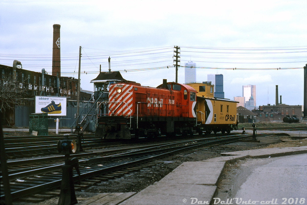 Relics of the past: CP S2 7026 and van 437227 amble over Strachan Avenue grade crossing, passing the crossing tower as they head north into Parkdale Yard as light power. At the time a gateman manning the crossing tower still manually operated the Strachan Ave. gates and warning lights for the CN Weston Sub and CP Galt Sub that crossed here on their way to the Toronto Terminals Railway territory downtown (the green shack near the Adidas sneaker ad was apparently the gateman's loo). Coloured switch lanterns mounted on switchstands dot the south end of Parkdale Yard's tracks. Visible in the background are the old John B. Smith & Sons building on the left (a former lumber dealer), the city's Wellington Destructor (garbage incinerator) on the right, and two tank cars spotted on the siding for the Toronto Municipal Abbatoir / Quality Packers ("tank siding", off of CN's Weston Sub). Modern office towers in the distance contrast with the gritty blue-collar industrial environs of the Parkdale area.

One of many small switchers built by Alco (and later MLW) for CP, the little 1000hp 7026 dates back to 1945 when CP was still ordering brand new steam engines (for road service) alongside diesel yard switchers. By 1979, the motive power assignment sheets show 7026 was one of nearly 30 little S2, S3 and S4 switchers assigned and maintained out of John St. Roundhouse downtown for local and yard switching duties in the Toronto area out of CP's various yards (Parkdale, Lambton/West Toronto, etc).

CP was also still using ancient steam-era wooden vans in local and transfer service in the 1980's. 437227 is notable in that it still has its old tounge-and-groove siding (escaping the plywood treatment most wooden vans got) and has its multimarks applied on the cupola end, where typical CP practice saw it almost always had it applied on the non-cupola end of wooden vans.

In less than a decade, declining industrial manufacturing in the areas surrounding downtown Toronto would result in both Parkdale Yard and John St. Roundhouse closing, and  with it the fleet of remaining steam-era Alco/MLW diesel switchers (replaced by more modern rebuilt switcher units). The abolishment of vans on freights in the late 80's also caused the newer modern Angus-built steel vans that had been in mainline run-through service bump the older steel and wooden vans from local service into retirement. Today aside from a few remaining industrial buildings (John B. Smith and the Wellington Destructor) this area is almost unrecognizable as modern condos have arisen in Libery Village and King West Village on many of the former industrial lands, and the crossing at Strachan has been eliminated in favour of a giant grade separation ducking under the roadway.

Bill McArthur photo, Dan Dell'Unto collection slide.

For a view of the area shot from Strachan Avenue a few years prior to the grade separation, see: http://www.railpictures.ca/?attachment_id=15195.