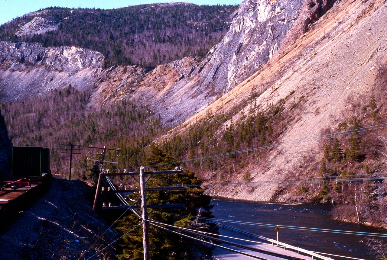HUMBER VALLEY VISTA. The incredible engineering feat of building a railway through this narrow section of the Humber Valley is evident in this scene taken from Coach 757 of Terra Transport's Extra 945 West on April 20, 1987. As the narrow gauge train snakes around the bend, the churning waters of one of the Island's most famous salmon rivers, the Humber, rush by Shellbird Island on the bottom right. Said to contain hidden pirate's treasure, this area was explored and charted by Captain James Cook some 225 years earlier with the railway going through around 1896/97. A favourite pastime in this area is locating the 'Man in the Mountain' a natural rock formation in the shape of a face on the upper right that looks down upon the the little island. With just a few miles to go, the photographer and his girlfriend's 138 mile journey (and first time on a Newfoundland train) from Bishop's Falls would soon come to an end in Corner Brook.