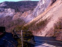 HUMBER VALLEY VISTA. The incredible engineering feat of building a railway through this narrow section of the Humber Valley is evident in this scene taken from Coach 757 of Terra Transport's Extra 945 West on April 20, 1987. As the narrow gauge train snakes around the bend, the churning waters of one of the Island's most famous salmon rivers, the Humber, rush by Shellbird Island on the bottom right. Said to contain hidden pirate's treasure, this area was explored and charted by Captain James Cook some 225 years earlier with the railway going through around 1896/97. A favourite pastime in this area is locating the 'Man in the Mountain' a natural rock formation in the shape of a face on the upper right that looks down upon the the little island. With just a few miles to go, the photographer and his girlfriend's 138 mile journey (and first time on a Newfoundland train) from Bishop's Falls would soon come to an end in Corner Brook.   