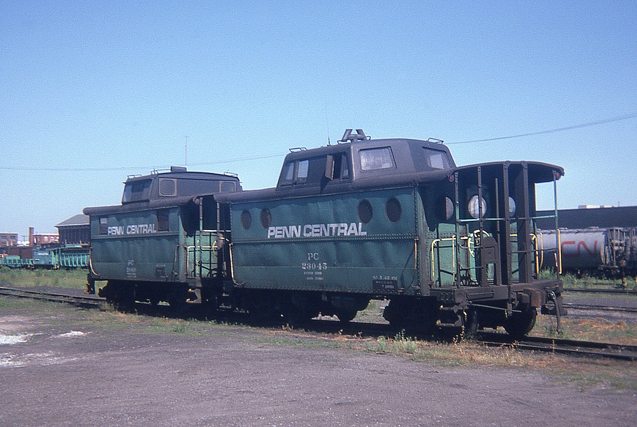 Two style examples of the old steel Penn Central cabooses sit out in the sunshine in downtown St. Thomas back in June of 1974. For location purposes note part of the huge station visible on the left background. I was unable to read the number of the furthest caboose.