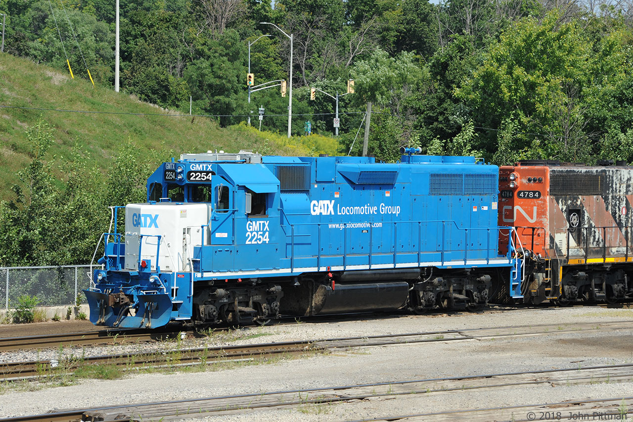 Remanufactured GP38-2 GMTX 2254 in fresh paint is paired with CN 4784, a GP38-2W. They are paused in between switching moves in Aldershot Yard, near Waterdown Road bridge. Behind them is the roadway providing access to Aldershot GO/VIA Station, its north parking lot, and Hwy 403 east.
A web search indicated that GMTX 2254 was originally an EMD GP38AC  built 11/1971 as blue GTW 5810, transferred to Central Vermont as CV 5810, then left the CN family. Said to have become LLPX 2217 (painted in UP colours).