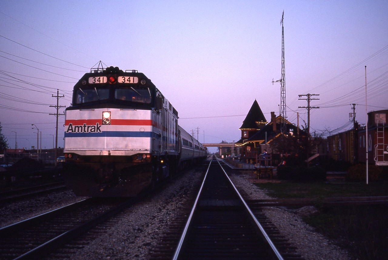 The new Amtrak "Maple Leaf" service from New York City to Toronto and return was only in its' 12th day of operation when I decided to wander down to Grimsby (I lived in St. Catharines at the time) to catch it at the station. What used to be known as Village Depot was no longer and I wanted to get a shot showing evidence of the old row of boxcars and the caboose at the near end on the right side of the photo before it all got hauled away. Grimsby station was home to a restaurant (Keystone Kellys) among other ventures back then when someone came up with the 'bright' (maybe 15 watt) idea of having a string of rail cars and having vendors sell out of each one. Sort of like a 'railroad plaza'. Well, the idea had its problems. For one thing it was very seasonal. And another, well, if there are no trains, where are the customers coming from? Boxcars sold trinkets, jewelry, souvenirs (!) ice cream and such. So, the great experiment didn't last long. Opened in 1977 and was sealed dead by 1979. Here it is spring 1981 and the cars and a lone caboose are still awaiting disposition.
Amtrak was a bit late on this evening, the Toronto-bound train arrived just before sunset. That was close.