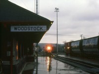 With the station's train order board semaphores set, the Woodstock station operator stands on the platform with his train order hoop at the ready and angled down at the rail, soon to be held up to "hoop up" train orders to the head end crew of an eastbound freight that's approaching the station on a gloomy evening just before 16:00 hours. Lead unit 5776 has its white classification lights illuminated, denoting this train as an extra freight. Such was common in the old days of train order territory operation when coloured flags, class lights, semaphores, train orders and precisely timed station clocks and pocket watches ensured the safe and timely movement of freight traffic.
<br><br>
<i>Keith Hansen photo, Dan Dell'Unto coll.</i>