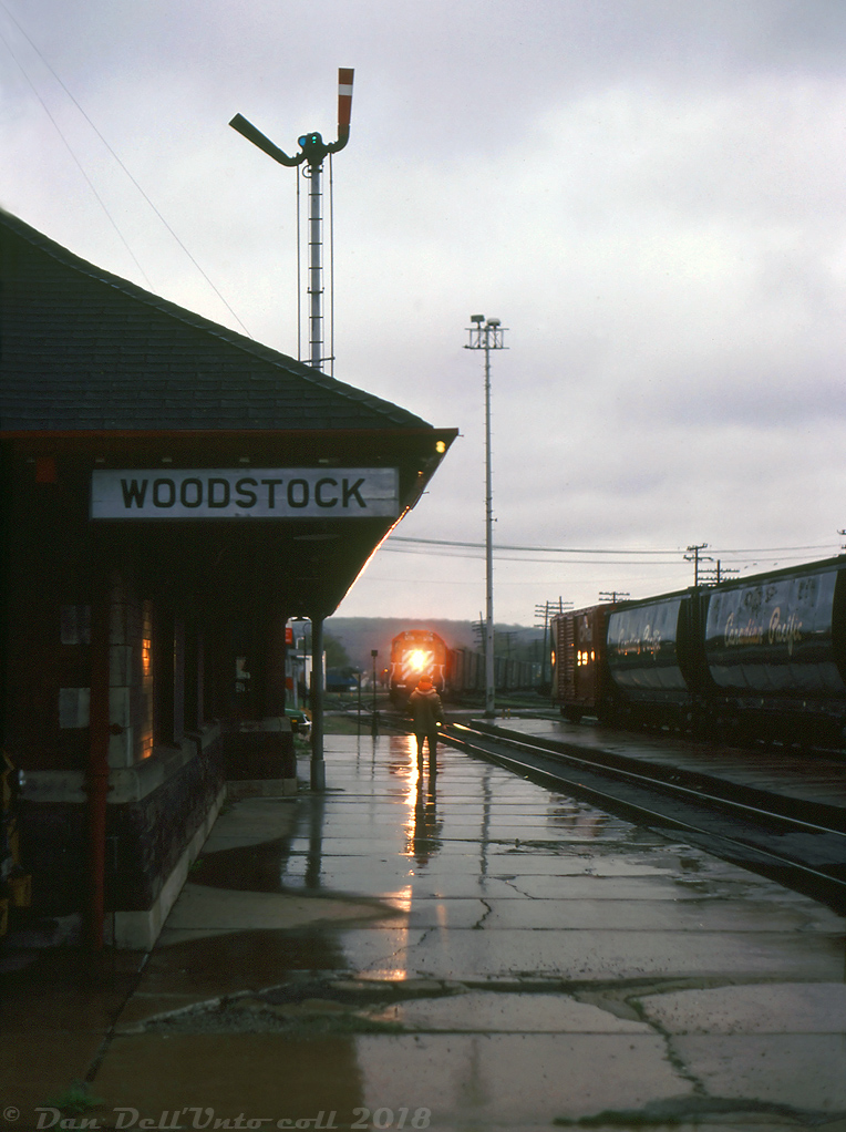 With the station's train order board semaphores set, the Woodstock station operator stands on the platform with his train order hoop at the ready and angled down at the rail, soon to be held up to "hoop up" train orders to the head end crew of an eastbound freight that's approaching the station on a gloomy evening just before 16:00 hours. Lead unit 5776 has its white classification lights illuminated, denoting this train as an extra freight. Such was common in the old days of train order territory operation when coloured flags, class lights, semaphores, train orders and precisely timed station clocks and pocket watches ensured the safe and timely movement of freight traffic.

Keith Hansen photo, Dan Dell'Unto coll.