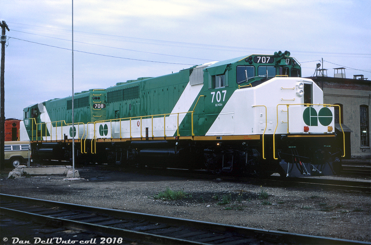 Freshly minted GO Transit GP40-2W's 707 and 708, recent graduates of the GMD's London locomotive plant, sit at CN's London Yard awaiting forwarding to their new owner. They were the first two units of GO's third -2W order being built (707-710, all shown as built October 1975) and were the only ones delivered new with 700-series numbers, unlike earlier orders that came as 9800's (renumbered to 700-706).

Rather than opt for steam-generator equipped units as they did with their GP40TC order, GO Transit in the 70's opted to expand its fleet with the standard freight-service GP40-2 model equipped with 76mph (60:17) gearing and the new CN-designed Canadian Safety Cab (note: officially there was no "W" in the designation, it was added by railfans). HEP-equipped cab cars rebuilt from old Ontario Northland F-units would be used with the freight GP's to provide electrical power for the passenger cars. It's interesting to note that GO Transit was the first buyer of the GP40-2 model equipped with the safety cab: GO 700-703 were ordered in December 1973 and built in January 1974, just before CN's first order of 9400's were built beginning in March 1974. However, by that time CN had already taken delivery of GP38-2W 5560 (4760) and 3500-series MLW M420's with the new cab design earlier on in 1973. 

GO 707, 708, and all but one of their sister units would eventually be displaced by new F59PH's and join the CN fleet in 1991, becoming 9674 and 9675 respectively.

Gord Taylor photo, Dan Dell'Unto collection.