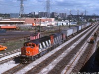 A short CN freight made up of piggyback traffic with GP40-2L(W) units 9585 and 9649 in the lead heads westbound on the Oakville Sub near Mimico East, about to pass under the busy Queen Elizabeth Way where it climbs up and over CN's main rail corridor from the west into downtown Toronto. A track gang is busy working on replacing sections of rail on the #4 main track, and the freight is on the #1 main - there were four mainlines numbered #1-4 from the north, plus a long service track and runaround track on the north side for local freights switching customers. Apartment buildings, the "green canopy" and housing in the background are from the Swansea and High Park neighbourhoods on the other side of the Humber River.<br><br>Back in the day there were still many online rail customers in the Swansea-Mimico area here, many involved in the food industry. On the left (out of frame) is the sprawling Ontario Food Terminal, that had at least ten rail sidings running into the property for delivering fresh produce and other perishables in reefers and insulated boxcars. Visible in the middle is a large warehouse belonging to the Oshawa Group (IGA, Food City, Towers, etc - note some of the trailers), which also had a pair of rail sidings to their building. In the far distance where the line curves towards Toronto, one can see part of the Stelco Swansea Works, also served by rail (established here in 1882 as the Dominion Bolt & Nut Co, near where the old Humber Belt Line spur ran). The famed Mr. Christie's plant was also a rail customer in the immediate area, although not visible here (behind the photographer on the other side of the QEW). Following years of closings and redevelopments, only the Ontario Food Terminal remains a going concern (and devoid of any rail service). Most of the rail traffic here today are GO and VIA trains, and one would be risking life and limb pulling over on this part of the busy Gardiner Expressway* trying to get this angle today!<br><br><i>Larry Moss photo, Dan Dell'Unto collection slide</i><br><br><i>(* Note: This part of the QEW was downloaded from the province to the city in 1997 and re-designated part of the Gardiner Expressway)</i>