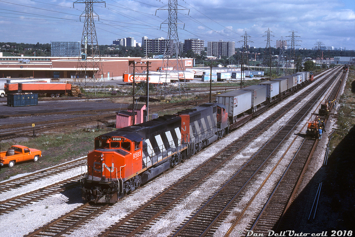 A short CN freight made up of piggyback traffic with GP40-2L(W) units 9585 and 9649 in the lead heads westbound on the Oakville Sub near Mimico East, about to pass under the busy Queen Elizabeth Way where it climbs up and over CN's main rail corridor from the west into downtown Toronto. A track gang is busy working on replacing sections of rail on the #4 main track, and the freight is on the #1 main - there were four mainlines numbered #1-4 from the north, plus a long service track and runaround track on the north side for local freights switching customers. Apartment buildings, the "green canopy" and housing in the background are from the Swansea and High Park neighbourhoods on the other side of the Humber River.Back in the day there were still many online rail customers in the Swansea-Mimico area here, many involved in the food industry. On the left (out of frame) is the sprawling Ontario Food Terminal, that had at least ten rail sidings running into the property for delivering fresh produce and other perishables in reefers and insulated boxcars. Visible in the middle is a large warehouse belonging to the Oshawa Group (IGA, Food City, Towers, etc - note some of the trailers), which also had a pair of rail sidings to their building. In the far distance where the line curves towards Toronto, one can see part of the Stelco Swansea Works, also served by rail (established here in 1882 as the Dominion Bolt & Nut Co, near where the old Humber Belt Line spur ran). The famed Mr. Christie's plant was also a rail customer in the immediate area, although not visible here (behind the photographer on the other side of the QEW). Following years of closings and redevelopments, only the Ontario Food Terminal remains a going concern (and devoid of any rail service). Most of the rail traffic here today are GO and VIA trains, and one would be risking life and limb pulling over on this part of the busy Gardiner Expressway* trying to get this angle today!Larry Moss photo, Dan Dell'Unto collection slide(* Note: This part of the QEW was downloaded from the province to the city in 1997 and re-designated part of the Gardiner Expressway)