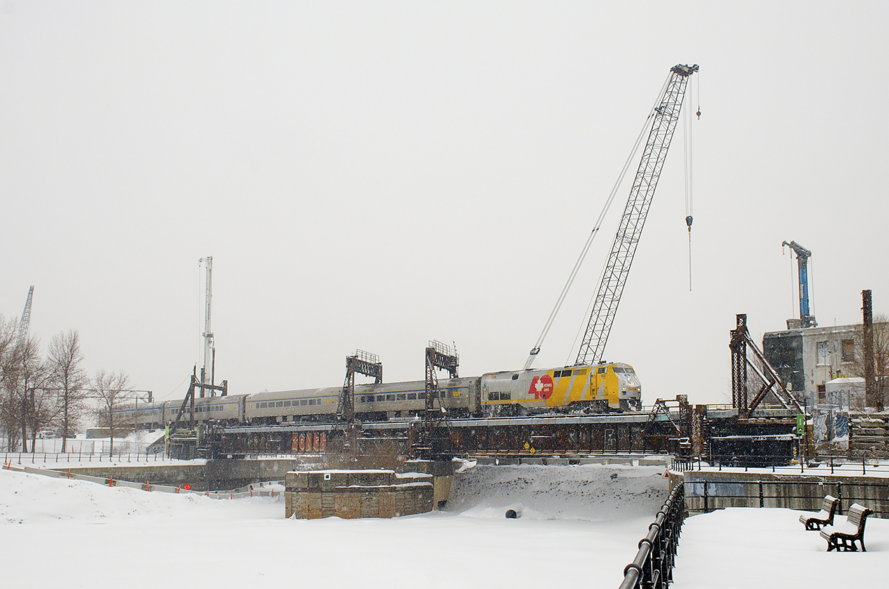 VIA 34 with VIA 920 and four HEP cars crosses the Lachine Canal on a snowy afternoon in Montreal. In the background, preparatory work for the REM (Réseau express métropolitain) light rail project has begun.