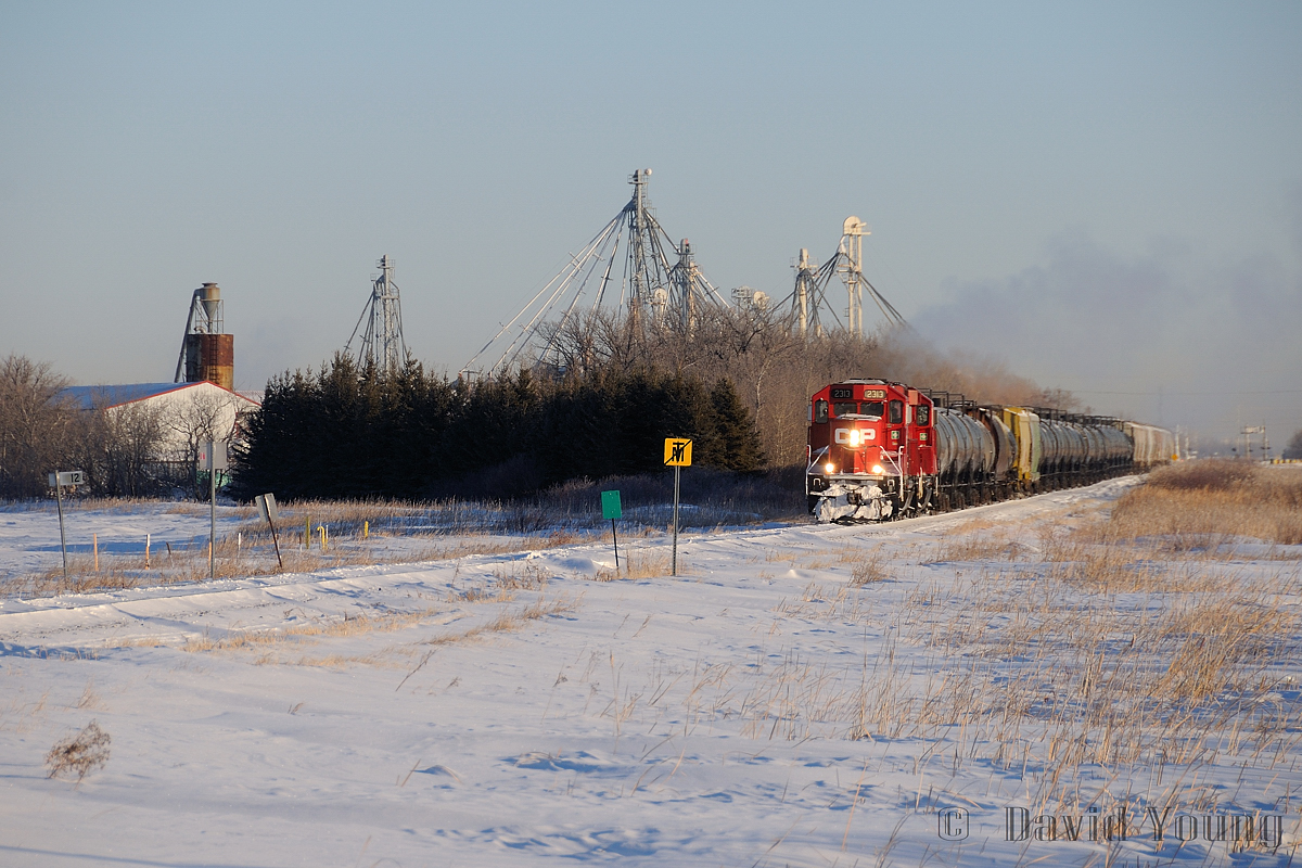 With OCS clearance in hand from Mile 12 to Gretna Spur Track Switch with a protect against one foreman, "The Altona", CP 2313 West rolls due south (timetable west), clearing the "Perimeter Highway" enroute to Altona, Manitoba. On a clear prairie day where the mercury is hovering around -30C a trail of exhaust hangs in the air as the crew navigate the last few miles of non-Main Track territory. Shortly they would be notching up to track speed (30 MPH), however the Rule 43 green flag protecting a slow order right at the begin/end main track will hamper their efforts for getting up to track speed.
