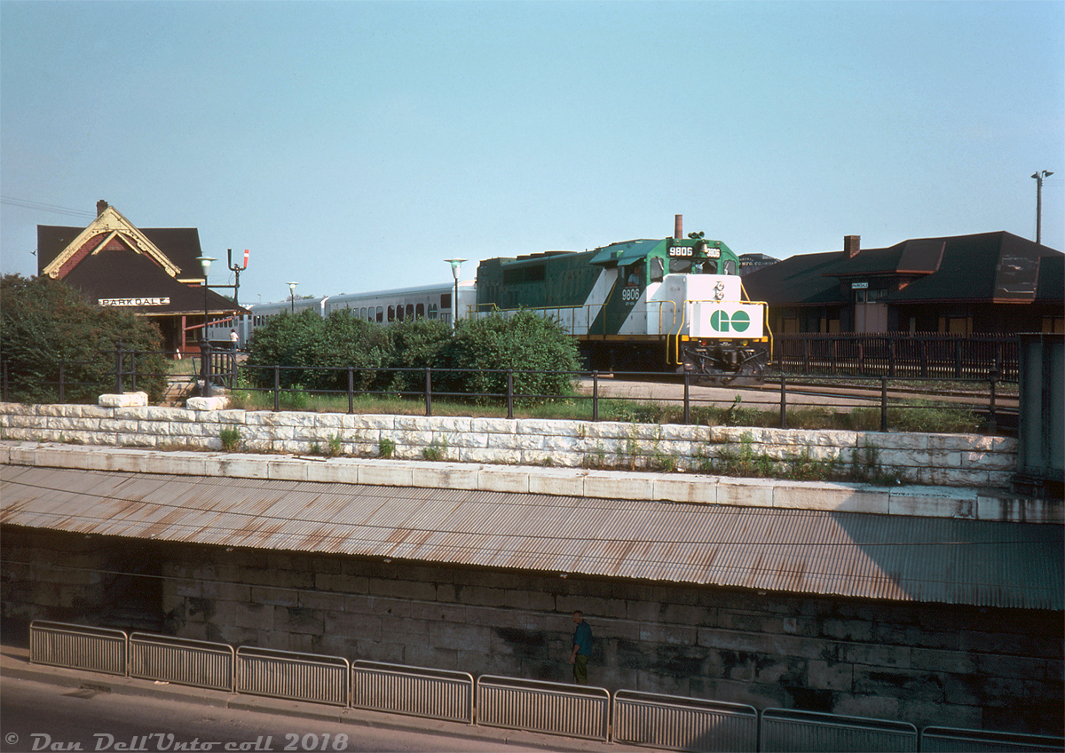GO Transit GP40TC 9806, freshly rebuilt by CN at Pointe St. Charles shop in late 1974 and repainted in the new green and white livery, is seen pulling a train of GO single-level Hawker Siddeley cars westbound on the Weston Sub past both CN's Parkdale Station and CP's Parkdale Station. The busy Queen Street underpass (subway) is visible in the foreground - note the cutout in the stone retaining wall that lead to a stairway up from sidewalk to platform level. Freight yards belonging to both railways were also located here at the time, along with numerous industries and sidings that still received rail service.According to another slide I have, this was shot right after a short CN passenger train (likely #161, CN still ran some passenger services up the Weston & Newmarket Subs but the only ones scheduled to stop at Parkdale were 160 & 161) had made its station stop at Parkdale, so going by timetables this is likely evening GO train #997 seen at around 5:43pm. GO's Georgetown line began service the previous year but Parkdale Station was never a stop for any GO trains (GO built their own station to the north at Bloor Street), so this GO could be either passing by or held up waiting for a light (perhaps the CN train was held up ahead at the diamond for a CP freight, or it was stopped at CN's West Toronto station). Despite this, Parkdale was a junction point between the Weston & Newmarket Subs so scheduled GO train times by the station were still listed in CN's employee timetables.Both Parkdale stations had seen better times at this point, and were in their final years of existence: CNR's Parkdale station (once known as "North Parkdale") was built in 1878 by the Northern Railway and expanded over the years. When closed the next year in 1976, CN wanted to demolish it (of course) but through fundraising by the Parkdale Save Our Station Committee the station was moved to Sunnyside in early 1977. Unfortunately a fire was started inside by vagabonds, and what was left was demolished that October.CPR's Parkdale station (located directly across from CN's on the Galt Sub) was originally built in 1910, closed in 1968, and is seen here boarded up and out of use. It was finally demolished sometime in the late 1970's. It also had a stairway leading up to it, via an arch cut into the retaining wall off Dufferin Street leading up to platform level.Photographer unknown (possibly a Charles Begg slide), Dan Dell'Unto collection.