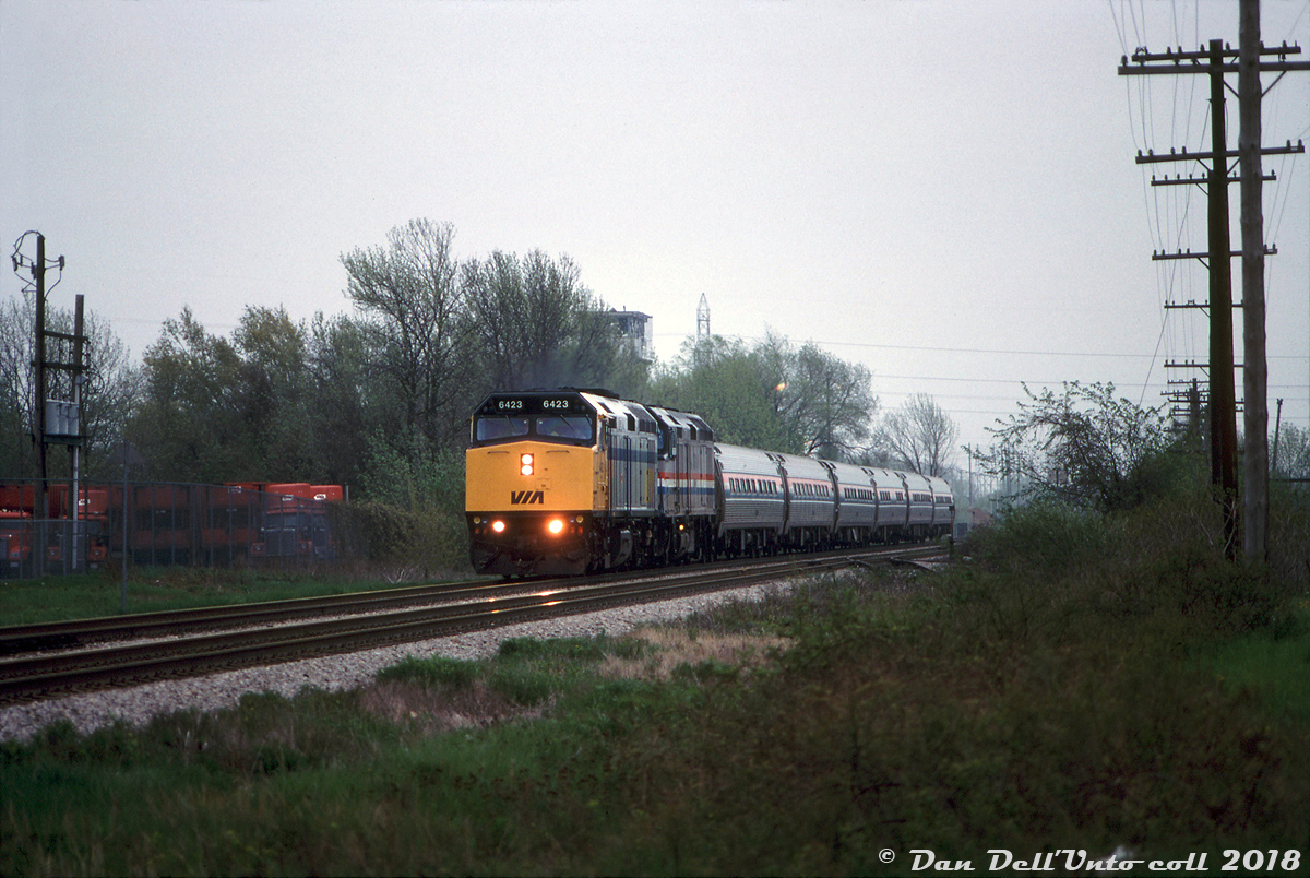 A gloomy Saturday evening finds VIA train #99, the US-Canada "Maple Leaf", heading westbound through Stoney Creek bound for Hamilton and Toronto. VIA F40PH-2D 6423 leads an Amtrak F40 (likely 296) and six Amfleet cars, passing by the old Waxman's scrap yard tower just west of SNS Stoney Creek where the Beach Sub once came off the Grimsby Sub near the hydro corridor. This was most likely taken from behind one of the lots off Cascade Street along the tracks, and if running on time would have been around 7:22pm going by the timetable. It's important to note that due to directional running at the time, this train was known as westbound #99 on the Grimsby Sub, and became #98 at Hamilton for running east to Toronto on the Oakville Sub (similarly, #97 from Toronto became #96 for Niagara Falls at Hamilton).Much thanks to Mr. Mooney for helping pinpoint where this was shot from, and helping sort out the power (Bill had this one mislabeled as the earlier eastbound #97, which had run with VIA 6423 and Amtrak 348. A VIA unit was apparently used to assist a few trains around that time, and would often be turned in the 'falls).Bill McArthur photo, Dan Dell'Unto collection.