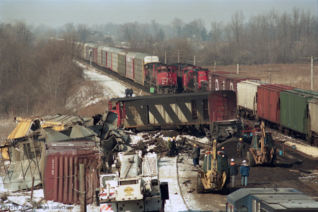 Excerpt from TSB Report - http://www.bst-tsb.gc.ca/eng/rapports-reports/rail/1995/r95s0021/r95s0021.asp 
On 16 February 1995, at 0349 eastern standard time (EST), a Canadian National (CN) freight train, travelling eastward on tangent main track at Mile 4.2 of the Strathroy Subdivision in London, Ontario, collided with the rear of a stationary freight train. The force of the collision propelled the stationary train eastward where it collided with another stationary freight train. Two employees sustained serious injuries. 
In this picture, apparently looking west from Hyde Park Road overpass, cleanup is well underway. 
Bombardier HR616 CN 2105, first unit into the crash, is off the rails, badly damaged, and was scrapped. 
The cab roof of GE Dash 8-40W LMSX 723 is at the bottom right - its rear hood etc looked pretty bad, but found  enough photos of LMS 723 at later dates to suggest that it was repaired and returned to service (or cloned). 
An eastbound train lead by CN 9596 is waiting, needing to pass close to the tail of CN 2105 on the repaired south track.  A westbound train on the north track is in motion past the site.
You can cut and paste the TSB report url above into your web browser - the full report is interesting. Don't know whether this scene was on the day of the crash, or a day or few after.