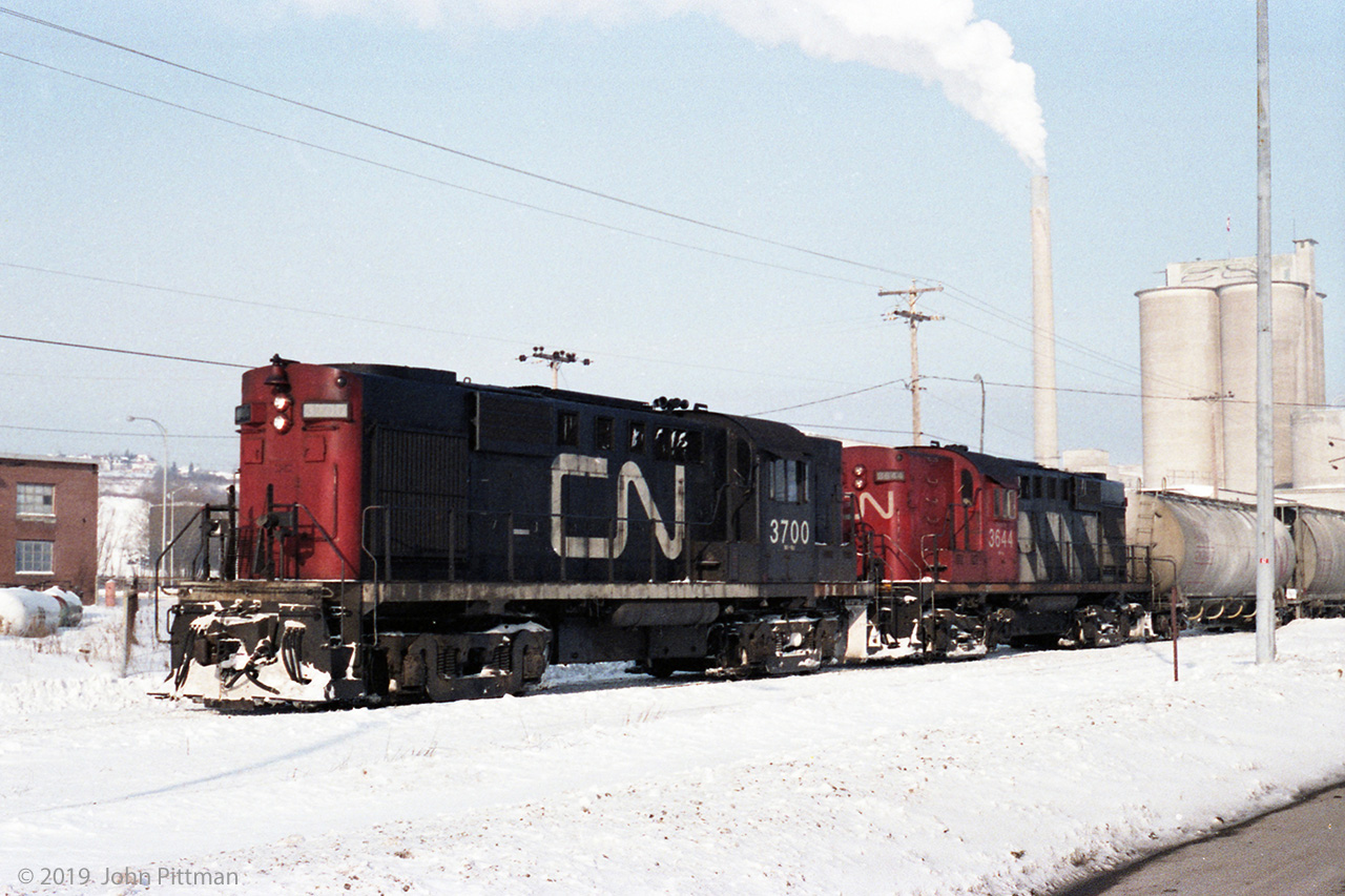 MLW RS-18's CN 3700 and CN 3644 were stopped with a train of cylindrical covered hoppers east of Quebec City on CN's spur along the north shore of the St Lawrence River, in the vicinity of Montmorency Falls.  The hoppers could be carrying cement, or maybe inputs for cement making, as the nearby industry looks to me like a cement manufacturing facility.  
As of 2019, could not find any trace of this plant using mapping resources - my conclusion is that it has been demolished.