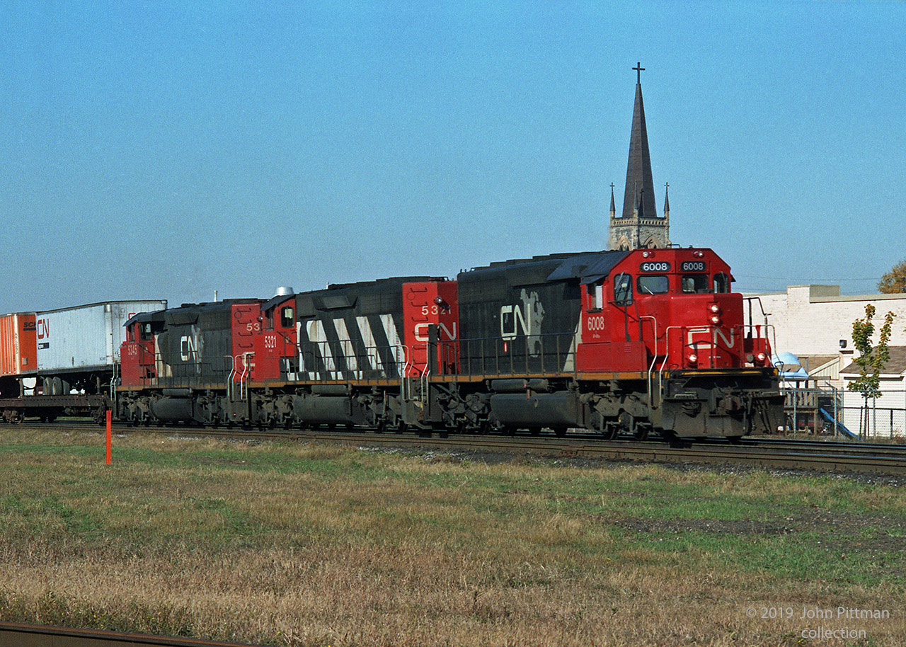 A CN eastbound with piggyback highway trailers (TOFC) on the front end is approaching Rectory Street grade crossing in London. The spire of St. Mary's Parish RC church crowns the first of three SD40-2 variants. CN 6018 is an SD40-2Q remanufactured from CN 5166, in its original CN North America paint scheme, while the others two are SD40-2w. 
First trailer says "CN Piggyback" - don't know what percent of the train was piggybacks.  
With some detective work I figured out the location and church. The date is more elusive. References indicated CN 6000-6028 remanufacture program was in 1993, while CN North America began mid-1992.  If anyone knows when CN 5345 got its CNNA repaint, when CN stopped Piggyback services through London, or the train number, please let us know in a comment.