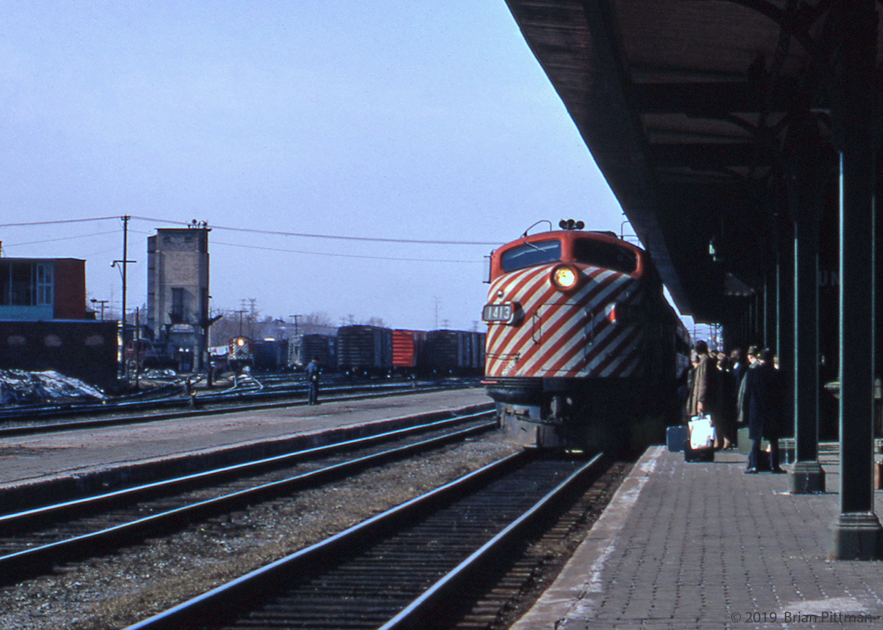 A westbound train from Quebec City powered by GMD FP9A CP 1413 is arriving at Trois-Rivieres station, with passengers travelling to Montreal on the platform to board. Over to the left (east) is the freight yard with boxcars predominant. The grey coaling tower has a continuing purpose as a high place for yard lighting. MLW RS-10 CP 8561 in CP Rail paint can be seen to the right of the tower working the yard, while a maroon and grey RS-23 switcher is to its left. A yard worker is standing on the between-tracks platform; he could be figuring out CP 8561's next switching moves. The building at left across the tracks masks the yard office and roundhouse beyond it; a pile of winter snow lingers beside it. Caboose track is left of that building, just out of the picture.
Scan and caption by John Pittman.