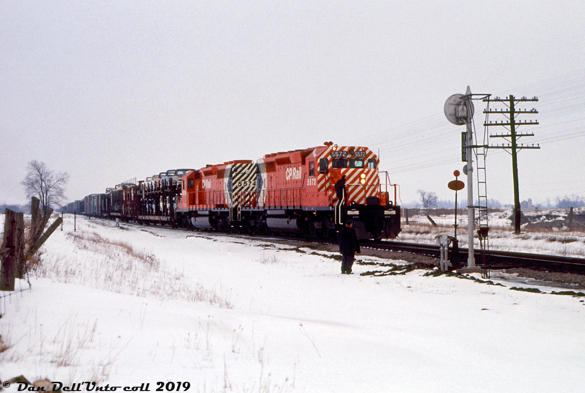 A drab winter's day is brightened up by a pair of shiny new SD40-2's, CP 5572 and 5574, working an westbound freight coming out of the west siding switch at Nissouri onto the Galt Sub, presumably after a meet with an eastbound. The white flags and class lights suggest this is an extra that would take the siding for higher class freights during meets, despite westbound being the superior direction. A cut of open autoracks full of new pickups and autos follow at the head end, and switch brooms stand at the ready in case the crew needs to sweep snow clear from the switchpoints. The Mile 104.9 marker plate on the signal suggests this is just east of Purple Hill Road., before the siding was extended westward at a later date (expanded from 6350 feet to 10150 feet, sometime in the 90's or 00's).At the time this photo was taken, CP was taking delivery of its first order of the new and improved "Dash Two" SD40-2 model, and the Dean & Hanna CPR book lists both 5572 and 5574 as outshopped from nearby GMD London and added to the roster on the same date: February 26th 1972 (the 6th and 7th SD40-2's received). So even if this shot of both working together wasn't their maiden run, it couldn't have been taken too long after (the slide is dated March 1972). Ironically, both were retired at the same time (in September 2004) and sent via NRE to work in Brazil, becoming América Latina Logística 9457 and Ferrovia Centro Atlantico 4815 respectively. A warm retirement career and a long way from the frigid Ontario winters they started working in.Gord Taylor photo, Dan Dell'Unto collection slide
