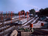 Twelve brand new H1 subway cars, part of the 164 car order built by Hawker Siddeley Canada for the new Bloor-Danforth subway line (opening early next year) sit together at the south end of the TTC's new Greenwood Subway yards on a Summer evening in July of 1965. The TTC had been taking delivery of the new cars from HSC's old Canadian Car & Foundry plant in Thunder Bay via flatcar since May, starting with the first two cars 5336 and 5337 delivered to <a href=http://www.railpictures.ca/?attachment_id=32616><b>Davisville Yard</b></a> while Greenwood Yard was still under construction, and the rest of the order to 5499 via Greenwood - seen here up to the 5360-series deliveries. Greenwood was constructed in the early-mid 60's in conjunction with construction of the new B-D subway line, in order to service and store the large fleet needed to operate the new east-west route. <br><br> Noted in the yard in the foreground are H1 cars 5361 & 5362 (not the proper final mated pair*). Lined up on the left is home-made tunnel clearance car RT-11, and H1's 5367-66-69-68-57-60 grouped together. The two car pair near the back of the yard is 5349-48. Two new deliveries from CN are also visible: 5363 riding on a CP 313000-series flatcar in the background waiting for her turn to be spotted and unloaded, and 5364 on another flatcar spotted in the "pit" ready for unloading. The pit track consisted of the railway-gauge siding track dead-ending in a depressed area, with a section of TTC-gauge track running level with the flatcar deck for wheeling subway cars off and into the yard using a car mover/trackmobile. There was another railway-gauge track nearby, the far right track, that had an overlapping dual-gauge section (perhaps for delivery of track supplies into the yard). New subway cars were delivered into the property via a railway spur running off CN's Kingston Sub into the <a href=http://www.railpictures.ca/?attachment_id=30975><b>back of Greenwood</b></a>. <br><br> At this time Greenwood was busy taking delivery of new cars, but still under construction and not yet fully operational (yard tracks were still being laid when the first two H1's were delivered to Davisville in early May 1965). Most of the track appears in place now, but one can see dug-up trenches in the yard, reels holding cable to be laid, fresh ballast spread around and piles stockpiled on the far right. The bright red elevated platforms are fire towers for easier access to any fires or incidents that may arise in the yard, a feature <a href=http://www.railpictures.ca/?attachment_id=818><b>still present today</b></a>. And the three smokestacks in the background mark the Richard L. Hearn Generation Station in the Toronto Portlands.<br><br> <i>John F. Bromley photo, Dan Dell'Unto collection slide scan. <br><br> *(Note: proper mated "A + B" pairs would be cars with numbers ending in 0-1, 2-3, 4-5, 6-7 and 8-9 mated together. Thus, 5360-5361, 5362-5363, etc).</i>