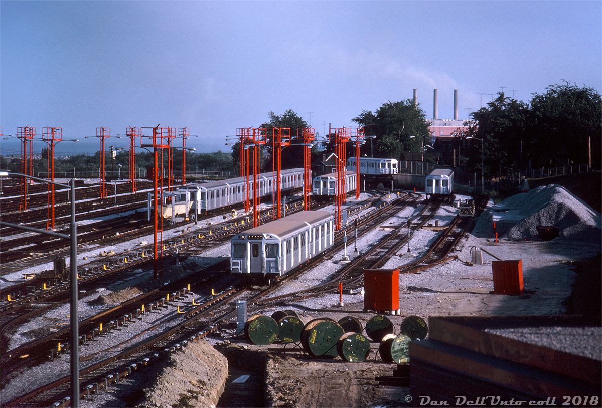 Twelve brand new H1 subway cars, part of the 164 car order built by Hawker Siddeley Canada for the new Bloor-Danforth subway line (opening early next year) sit together at the south end of the TTC's new Greenwood Subway yards on a Summer evening in July of 1965. The TTC had been taking delivery of the new cars from HSC's old Canadian Car & Foundry plant in Thunder Bay via flatcar since May, starting with the first two cars 5336 and 5337 delivered to Davisville Yard while Greenwood Yard was still under construction, and the rest of the order to 5499 via Greenwood - seen here up to the 5360-series deliveries. Greenwood was constructed in the early-mid 60's in conjunction with construction of the new B-D subway line, in order to service and store the large fleet needed to operate the new east-west route.  Noted in the yard in the foreground are H1 cars 5361 & 5362 (not the proper final mated pair*). Lined up on the left is home-made tunnel clearance car RT-11, and H1's 5367-66-69-68-57-60 grouped together. The two car pair near the back of the yard is 5349-48. Two new deliveries from CN are also visible: 5363 riding on a CP 313000-series flatcar in the background waiting for her turn to be spotted and unloaded, and 5364 on another flatcar spotted in the "pit" ready for unloading. The pit track consisted of the railway-gauge siding track dead-ending in a depressed area, with a section of TTC-gauge track running level with the flatcar deck for wheeling subway cars off and into the yard using a car mover/trackmobile. There was another railway-gauge track nearby, the far right track, that had an overlapping dual-gauge section (perhaps for delivery of track supplies into the yard). New subway cars were delivered into the property via a railway spur running off CN's Kingston Sub into the back of Greenwood.  At this time Greenwood was busy taking delivery of new cars, but still under construction and not yet fully operational (yard tracks were still being laid when the first two H1's were delivered to Davisville in early May 1965). Most of the track appears in place now, but one can see dug-up trenches in the yard, reels holding cable to be laid, fresh ballast spread around and piles stockpiled on the far right. The bright red elevated platforms are fire towers for easier access to any fires or incidents that may arise in the yard, a feature still present today.  John F. Bromley photo, Dan Dell'Unto collection slide scan.  *(Note: proper mated "A + B" pairs would be cars with numbers ending in 0-1, 2-3, 4-5, 6-7 and 8-9 mated together. Thus, 5360-5361, 5362-5363, etc).