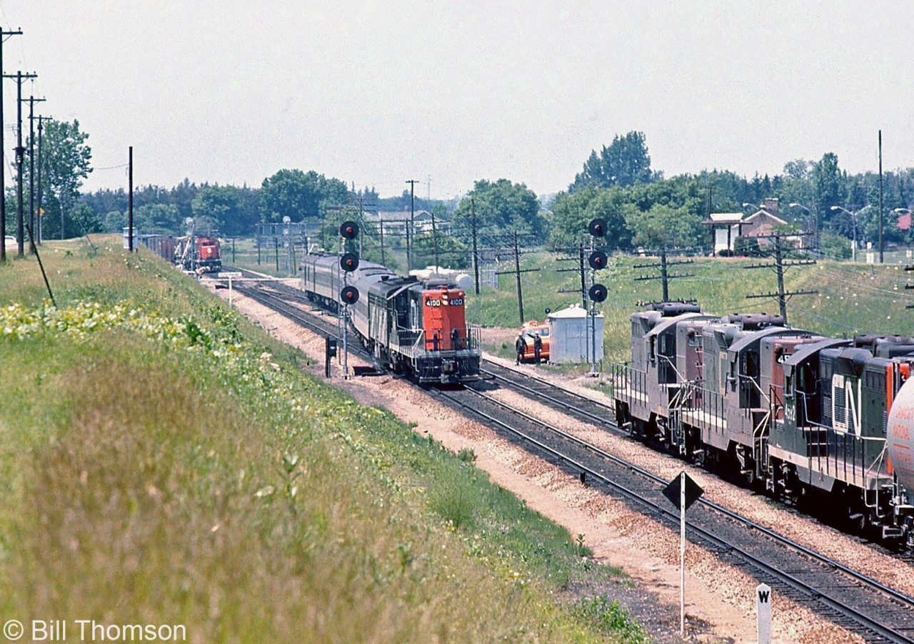 A three-way meet is shown at Hardy (just west of Brantford at Mile 24.9 CN Dundas Sub): CN GP9 4100 and an FPB4 head an eastbound passenger train crossing over from the north to the south track, as a westbound freight lead by three CN GP9's waits on the north track, and an eastbound freight (with what looks to be an SD40 leading) waits on the south track just past the crossing. A pair of workers watch the scene by their orange CN van parked near the signal bungalow.