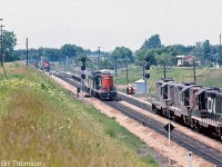 A three-way meet is shown at Hardy (just west of Brantford at Mile 24.9 CN Dundas Sub): CN GP9 4100 and an FPB4 head an eastbound passenger train crossing over from the north to the south track, as a westbound freight lead by three CN GP9's waits on the north track, and an eastbound freight (with what looks to be an SD40 leading) waits on the south track just past the crossing. A pair of workers watch the scene by their orange CN van parked near the signal bungalow.