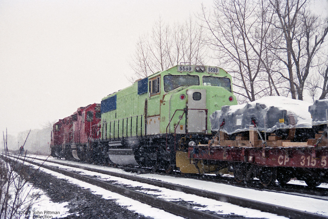 A snowy send off, with a CP Rail 8200 series GP9u and GP38-2 CP 3096 (last digit ?) leading a short train of new locomotives away from the GM Diesel Division Plant in London toward CP's Quebec Street Yard. At the switch ahead they will leave GM DD track and enter CP's Galt sub. 
The unit in primer is SD60i CR 5589, I guess Conrail paints it blue at Juniata Works in Altoona PA. 
On flat car CP 315502 are four 5' 3" gauge 3-axle trucks for Irish Rail (Iarnród Éireann) JT42HCW locomotives 229 and 231 on the last 2 flatcars of this train. Date provided based on approximate build dates and weather conditions.