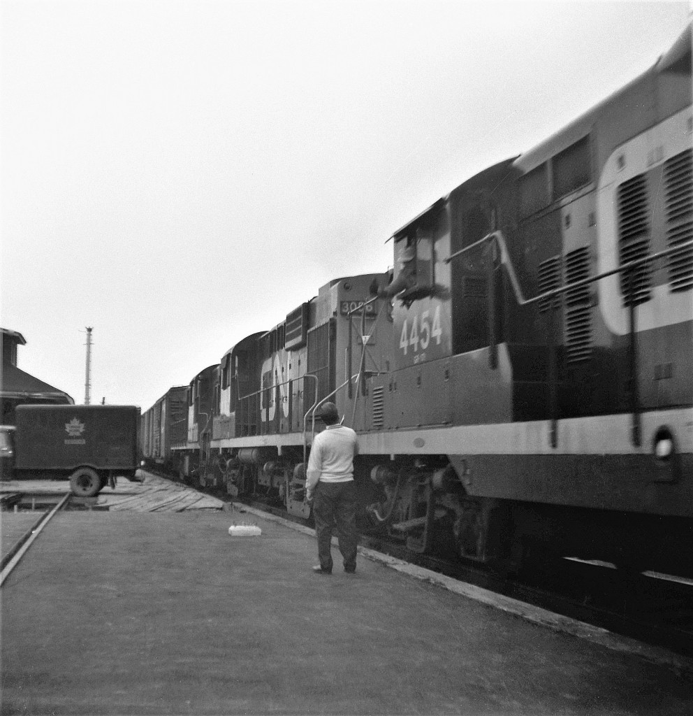 Early on Tuesday morning, August 31, 1965, CN train 401 lead by GP9 4454, RS-10 3086, and RS-18 38xx becomes the last train to pass the old wood frame Pembroke, Ontario station.  The station is visible behind the CN Express truck.  The operator, hooping up the orders to the engineer, moved into the new station the day prior. Work still continues on the platform.  Within one hour of the train's passing, the old station was bulldozed into a pile of rubble.  Location listed is a best guess on my part.