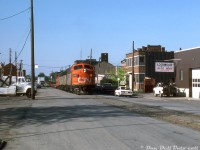 Matching CN zebra-striped A-B-A F-units 9173, 9195 and 9166 handle #725 "The Steel Train" along the street-running portion of CN's Hagersville Sub along Ferguson Avenue, having just departed Hamilton (Stuart St.) yard for their run up the escarpment to Caledonia and on to Nanticoke. The train is seen passing by the Lockwood Motors auto body repair shop, approaching Kelly St. after having just crossed Cannon St. moments before. Timetables show this section of the line was limited to a pokey 10mph, for obvious reasons due to the traffic and pedestrian hazards present. <br><br> As others have mentioned in previous photos, the Steel Train was one of the last holdouts to regularly use this street-running portion of the Hagersville Sub through the downtown streets of Hamilton (and only running empty up the escarpment; it returned loaded on the Dundas Sub via Brantford), but not for too much longer. After a bridge in Rymal was damaged around 1987(?), CN elected not to repair it and fully rerouted the steel train over the Dundas Sub. The line between Caledonia and Hamilton was severed into two spurs, operated as the Ferguson Ave Spur from Hamilton and Rymal Spur from Caledonia, but didn't fare much better due to low rail traffic. The revised (and present-day) version of the Hagersville Sub uses the former Dunnville Sub (from Brantford to Caledonia) to access the original portion between Caledonia to Nanticoke. <br><br> This would also be one of the last great hurrahs of CN's F-unit fleet (notably catching them regularly assigned in solid A-B-A sets), as the remaining units would soldier on for a few more years until they were retired in 1988-89 and sold off. <br><br> <i>Harold E. Brouse photo, Dan Dell'Unto collection slide.</i>