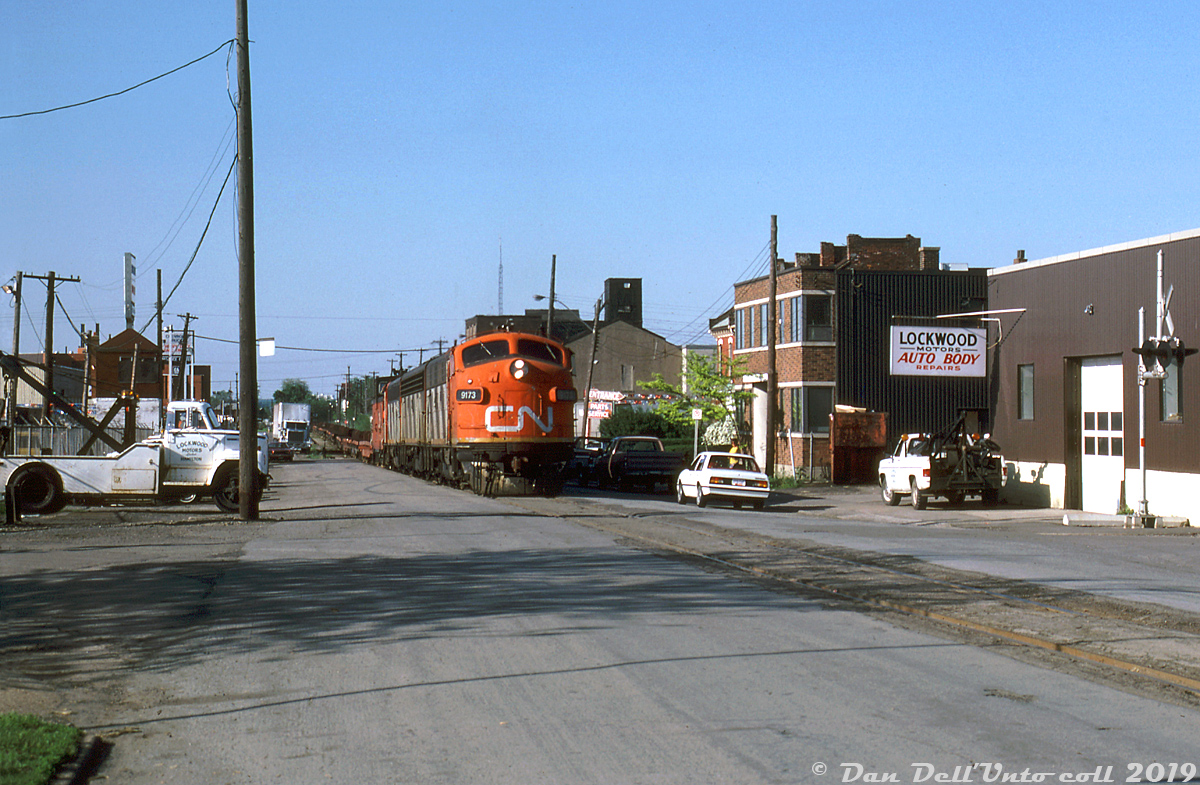 Matching CN zebra-striped A-B-A F-units 9173, 9195 and 9166 handle #725 "The Steel Train" along the street-running portion of CN's Hagersville Sub along Ferguson Avenue, having just departed Hamilton (Stuart St.) yard for their run up the escarpment to Caledonia and on to Nanticoke. The train is seen passing by the Lockwood Motors auto body repair shop, approaching Kelly St. after having just crossed Cannon St. moments before. Timetables show this section of the line was limited to a pokey 10mph, for obvious reasons due to the traffic and pedestrian hazards present.  As others have mentioned in previous photos, the Steel Train was one of the last holdouts to regularly use this street-running portion of the Hagersville Sub through the downtown streets of Hamilton (and only running empty up the escarpment; it returned loaded on the Dundas Sub via Brantford), but not for too much longer. After a bridge in Rymal was damaged around 1987(?), CN elected not to repair it and fully rerouted the steel train over the Dundas Sub. The line between Caledonia and Hamilton was severed into two spurs, operated as the Ferguson Ave Spur from Hamilton and Rymal Spur from Caledonia, but didn't fare much better due to low rail traffic. The revised (and present-day) version of the Hagersville Sub uses the former Dunnville Sub (from Brantford to Caledonia) to access the original portion between Caledonia to Nanticoke.  This would also be one of the last great hurrahs of CN's F-unit fleet (notably catching them regularly assigned in solid A-B-A sets), as the remaining units would soldier on for a few more years until they were retired in 1988-89 and sold off.  Harold E. Brouse photo, Dan Dell'Unto collection slide.
