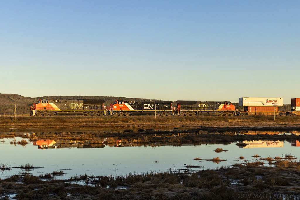 Shortly after the sun rises over the hillside, eastbound stack train Q120 comes rumbling by, giving off reflections at Upper Dorchester, New Brunswick.