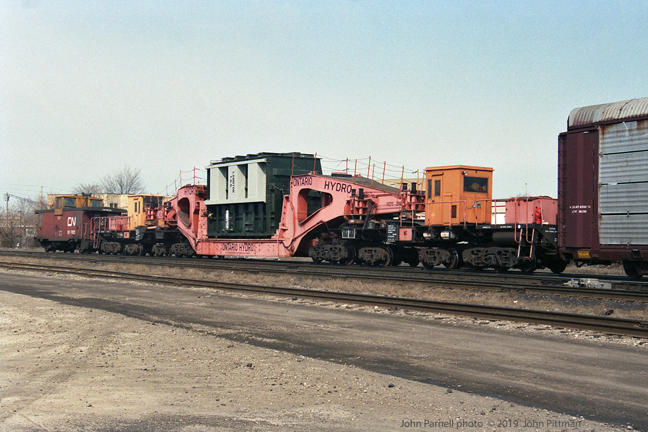 Schnabel car HEPX 200 has been in service for many years, moving heavy and oversize loads for Ontario Hydro and its successors. 
In this picture from 1992 it is lettered for Ontario Hydro with (looks-like) faded red paint, different from the orange Hydro One colour scheme it wears now. Another difference from recent pictures on RP.ca is that this load labelled "Lambton T.G.S." is carried on a cradle joining the car ends, rather than having the load slung between  the two ends of the Schnabel car.
From adjacent images, it appears that this train is eastbound at London Jct, branching off the Dundas Sub onto the Guelph sub. Other frames from the film with same date show GP9rm units CN 7029 (leading long hood forward) and CN 7039 - I believe they powered this train. The caboose is CN 78122.