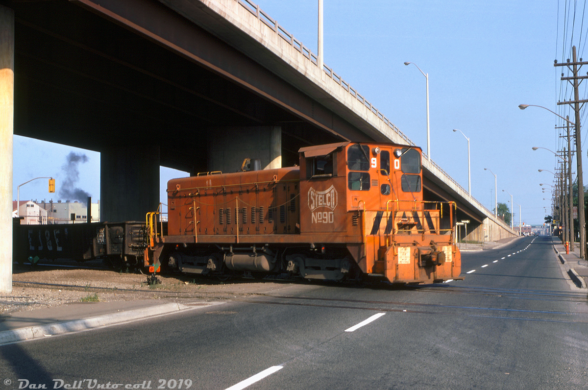 Stelco SW900 90 passes under the elevated portion of Burlington Street and crosses the street-level crossing just west of Wilcox Street, working outside Stelco's Hilton Works plant in the evening on the Hilton Works (J85) lead (that leads to the east side trackage). The track in the forground is the J80 lead that runs to the west side trackage in the plant. The unit is probably taking some outbound cars from Stelco to one of the nearby yards along the CN N&NW spur.  90 is one of a number of GMD SW8 and SW900 switchers built for Stelco in the 50's and 60's, featuring a special low-clearance cab and exhaust stacks for working in and around industrial areas. Originally built in January 1965 at GMD's London ON plant, at this point in time makeshift full handrails have been installed along the sides (chains and stanchions, also note the typical as-delivered hood-mounted handrails still present). As with other sister units, 90 has been ballasted heavier with large pieces of steel mounted to the end pilots, and a thin layer of steel added on top the frame walkways to bring total weight up to 137 tons (from around 115 tons a normal SW900 would weight). The unit sports the older pre-white safety stripes livery, and the old "dog bone" logo on the cab sides.  The number of Stelco's plant switchers has dwindled over time, as operations were decreased or rationalized some units were sold off or transfered to other operations, but a few remain at their Hamilton site for in-plant switching (including unit 90).   Original photographer unknown, Dan Dell'Unto collection slide.