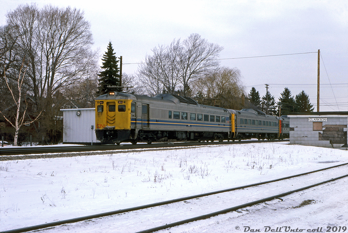 A train of VIA Rail Budd RDC's (in three different liveries: VIA-CN, VIA, and CN) head westbound through snowy Clarkson, passing the old CN Clarkson Station, long out of use and falling into disrepair. There would be no station stop here for the Budds today, nor has there been for nearly a decade.  This small brick single-story iteration of Clarkson Station was built by CN as a replacement for the previous GTR-built Clarkson Station that had burned down in 1962. But the new station would see only a few years of use, as regular passenger service here ended on Saturday May 20th 1967, the last day of CN's commuter train operations between Hamilton and Toronto (Mon-Sat CN trains #921 & #922 would be the final two to use the station, according to a 1965 timetable). Three days later, government startup commuter service operator GO Transit would start running trains on their Lakeshore corridor using the new Clarkson GO Station built to the west. As far as I have been able to find, CN's Clarkson Station building here met its eventual demise sometime between 1978 and 1984.  According to Wilfred Sergeant's "Building GO Transit" (2004) e-book, there were a number of decisions influencing this move. The CN station here was located in what once was the old "Clarkson's Corners" village and was constrained by established development and small local roads. However, land to the west along Southdown Road was vacant and presented the opportunity for better traffic flow via a grade separation, better traffic access off the main roads, and ample land for parking lots. CN's stations at Long Branch, Dixie Road, Lakeview and Lorne Park* would also suffer similar fates, either closed down and replaced with new GO stations in better locations, or consolidated with others nearby in order to achieve optimal station spacing for better train timing.  *Note, due to strong local community opposition, CN's Lorne Park station remained and had *one* GO train run scheduled to serve there each morning (GO #954 from Hamilton to Toronto, that would stop at Lorne Park instead of Clarkson at 7:58am each morning), until it was rescheduled to Clarkson after about a year.  Douglas J. Fear photo, Dan Dell'Unto collection.