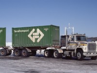 An A Train of semi-trailers bearing two 20' Terra Transport containers is seen in Johnstown, Ontario in April 1984.  The containers were new within the last five years with the advent of Terra Transport in September 1979.  Many of these containers lasted well beyond the end of the former Newfoundland Railway in September 1988.  A Ford tractor is in command at the Johnstown grain elevators.  For decades these elevators were the site of grain transshipments between 'Lakers' and 'Canallers.'  Prior to the opening of the Seaway in 1959, the small size of the locks between Cardinal, Ontario and Montreal precluded the use of 'Lakers' due to their larger size.  Winter storage continues to be a long-time feature of the elevators.  As well, boats were often wintered here when the lakes and Seaway were closed to shipping.  In the background, the M/V Cartiercliffe Hall is in winter layup.  The M/V Cartiercliffe Hall was built in Hamburg, Germany, in 1960 for service as an ocean ore carrier. In 1976 it was converted for use as a dry bulk carrier for Great Lakes service. In June 1979 downbound to Port Cartier from Lake Superior, it was involved in a fire in the crew quarters that killed seven of 23 crewmembers.  More details of the tragedy may be found here: http://continuouswave.com/misc/cartiercliffehall.html.  Sold and renamed AlgoOntario, the boat was sold for scrap in 2009.

The covered hoppers are on National Harbours Board tracks which were served by CN off the Kingston Sub and CP Rail off their Prescott Sub prior to its abandonment in 1997.  It was an interchange point between the railways and the switches were interlocked to prevent collisions.  