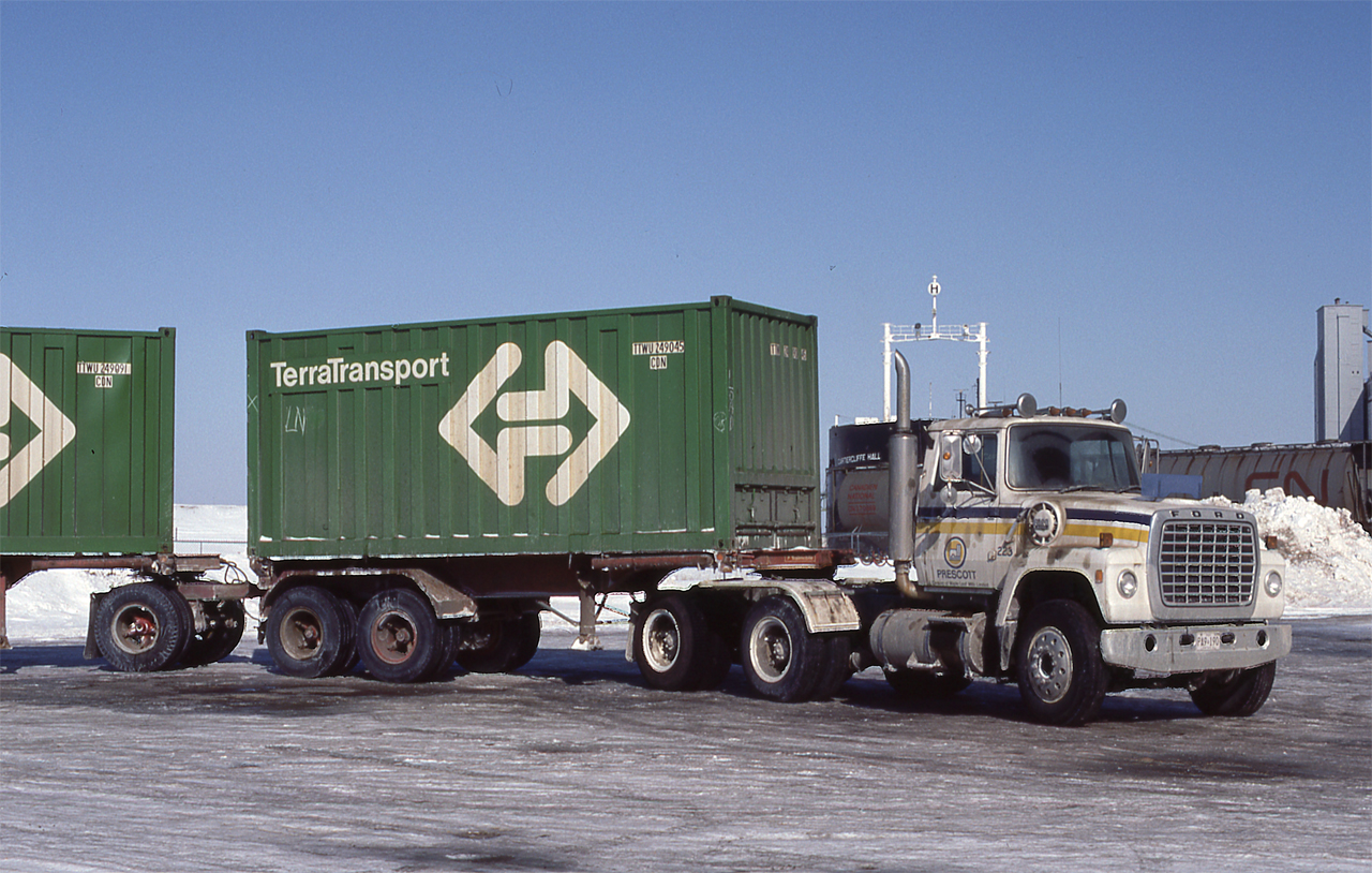 An A Train of semi-trailers bearing two 20' Terra Transport containers is seen in Johnstown, Ontario in April 1984.  The containers were new within the last five years with the advent of Terra Transport in September 1979.  Many of these containers lasted well beyond the end of the former Newfoundland Railway in September 1988.  A Ford tractor is in command at the Johnstown grain elevators.  For decades these elevators were the site of grain transshipments between 'Lakers' and 'Canallers.'  Prior to the opening of the Seaway in 1959, the small size of the locks between Cardinal, Ontario and Montreal precluded the use of 'Lakers' due to their larger size.  Winter storage continues to be a long-time feature of the elevators.  As well, boats were often wintered here when the lakes and Seaway were closed to shipping.  In the background, the M/V Cartiercliffe Hall is in winter layup.  The M/V Cartiercliffe Hall was built in Hamburg, Germany, in 1960 for service as an ocean ore carrier. In 1976 it was converted for use as a dry bulk carrier for Great Lakes service. In June 1979 downbound to Port Cartier from Lake Superior, it was involved in a fire in the crew quarters that killed seven of 23 crewmembers.  More details of the tragedy may be found here: http://continuouswave.com/misc/cartiercliffehall.html.  Sold and renamed AlgoOntario, the boat was sold for scrap in 2009.

The covered hoppers are on National Harbours Board tracks which were served by CN off the Kingston Sub and CP Rail off their Prescott Sub prior to its abandonment in 1997.  It was an interchange point between the railways and the switches were interlocked to prevent collisions.