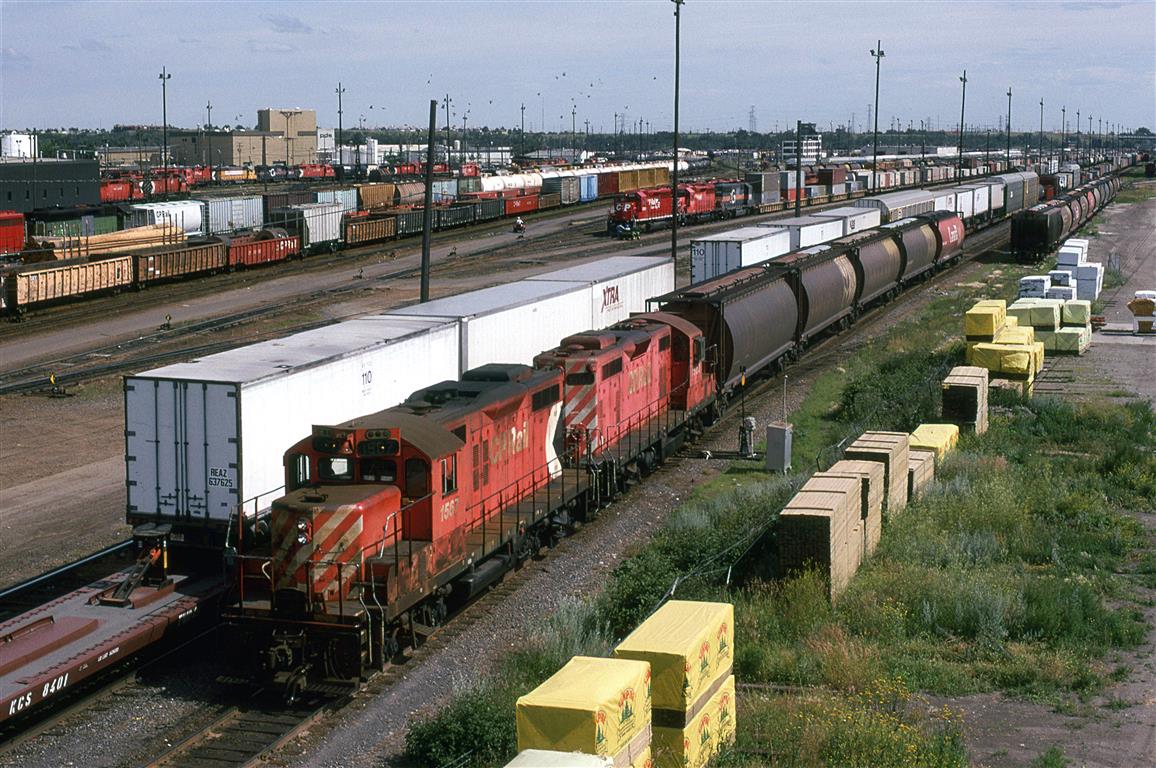 A yard job, or perhaps a local, has a string of 5 grain hoppers in tow, while a container train can be seen in the background.