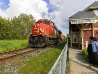 A young lady looks on as 3070 hustles train 305 by the old station at Riviere Bleue, PQ. 