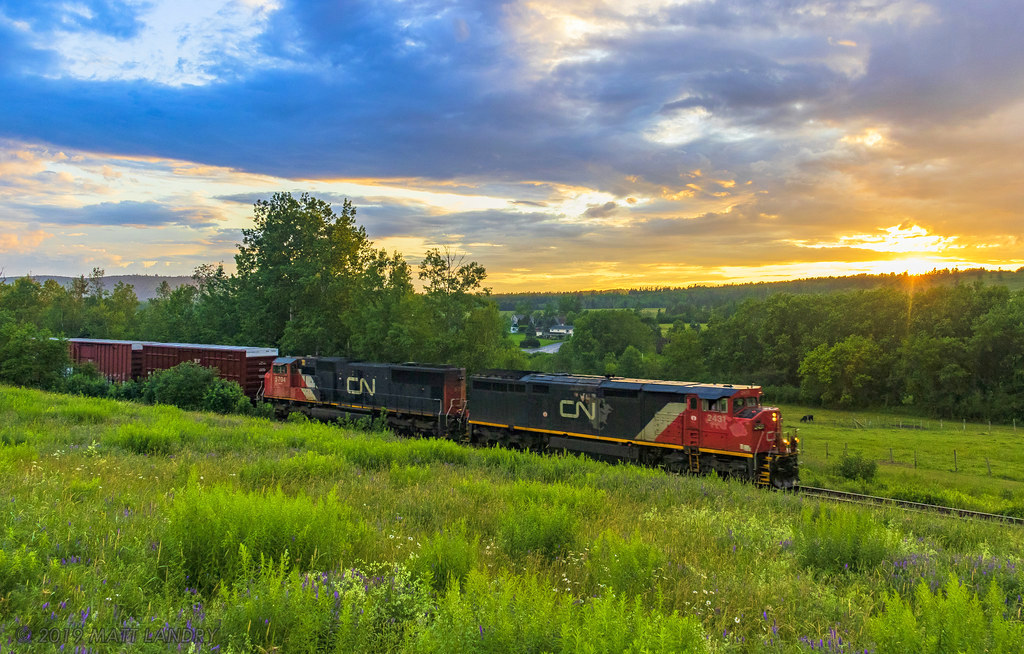 Nearing sunset, the sun dips below the clouds just enough to light up the sky and scene, as train 406 heads along the countryside, at Norton, New Brunswick.