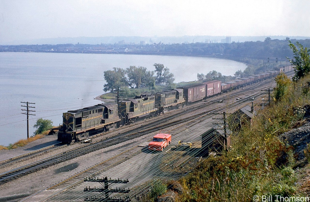 Canadian National RS18 3737 leads a GP9 and RS18 on an eastbound freight, curving around Burlington Bay on the Oakville Sub at Hamilton Junction, passing  by a red CN maintenance of way truck and some wayside section houses. Trailing behind the Santa Fe and Pennsylvania RR boxcars near the head end are a cut of loaded CN bottom-dump gondolas. In the background, one can make out CN's Hamilton (Stuart Street) yard and surrounding industries.