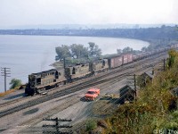 Canadian National RS18 3737 leads a GP9 and RS18 on an eastbound freight, curving around Burlington Bay on the Oakville Sub at Hamilton Junction, passing  by a red CN maintenance of way truck and some wayside section houses. Trailing behind the Santa Fe and Pennsylvania RR boxcars near the head end are a cut of loaded CN bottom-dump gondolas. In the background, one can make out CN's Hamilton (Stuart Street) yard and surrounding industries.