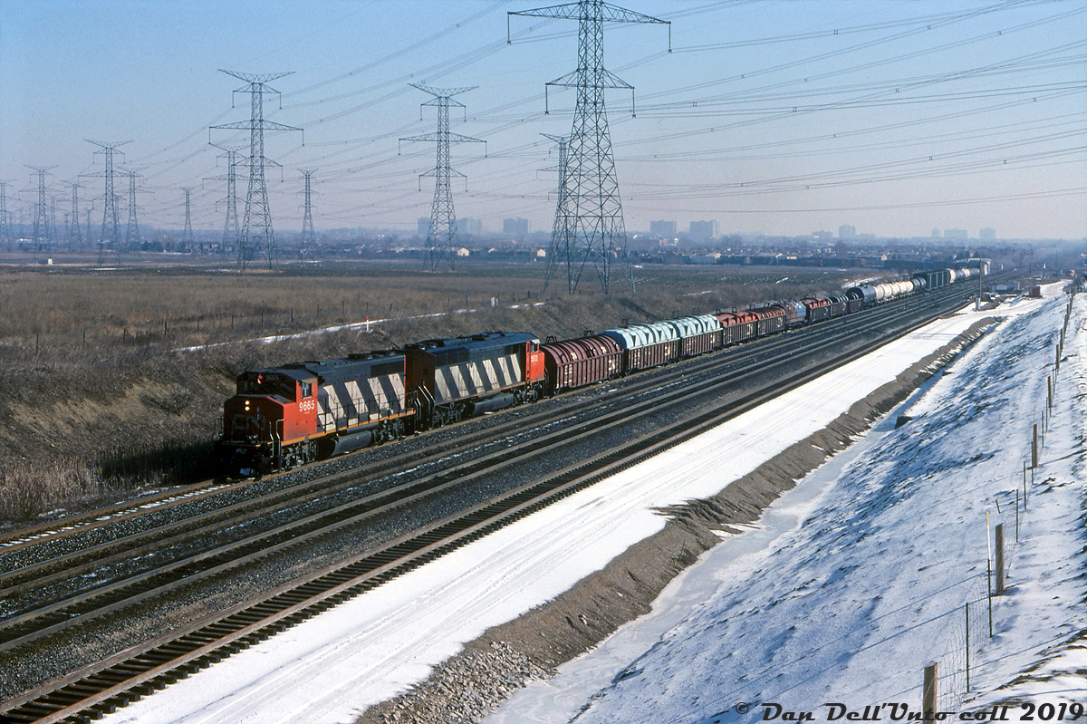 CN GP40-2W 9665 and SD50AF 5502 handle train #452, coming off the CN Newmarket Sub southbound and heading westbound on the York Sub at Snider, about to duck under Keele Street and enter CN's MacMillan Yard.The York Sub from Vaughan to Pickering Junction (and Halton Sub from here to Halwest) was built in the early-mid 60's as part of the "Toronto bypass" project to divert mainline freight traffic out of downtown Toronto and the surrounding urban areas with a new rail line built to the north, running to a new larger and more modern freight classification yard (CN Toronto Yard, later renamed MacMillan Yard) in Concord-Maple area of what is now the City of Vaughan. Since the north-west running Newmarket Sub had already existed here for decades (originally built by the Northern Railway of Canada in the 1850's), an interlocking diamond was required for the new York Sub to cross it, and the location named "Snider" (Snider was a long-established family name in the area, and they owned farms and mills in years past). Today, the Snider interlocking diamond shown in the background has been replaced with a large flyover in order to handle more frequent Barrie line GO train service.Karl Bury photo, Dan Dell'Unto collection.