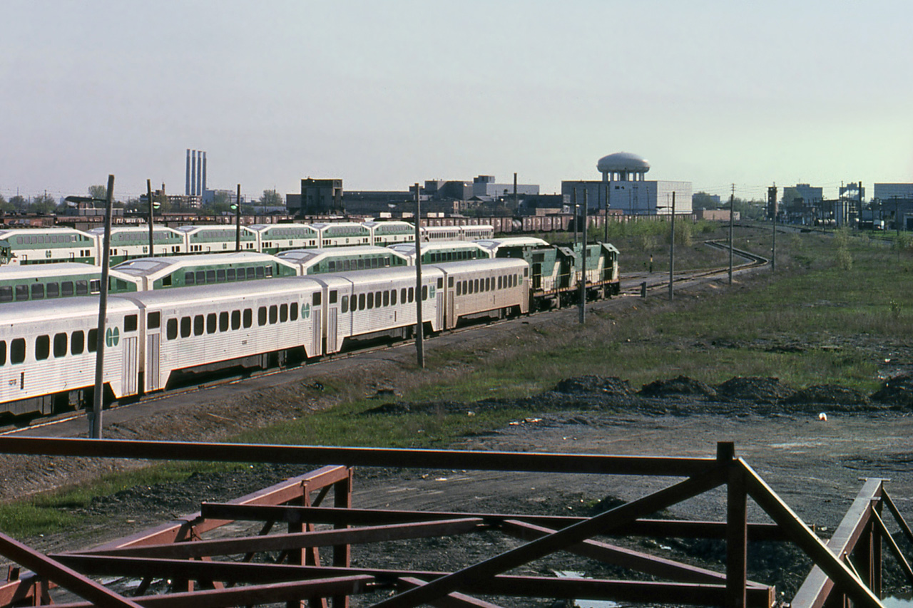 A mid-year afternoon weekend view of GO Transit's Willowbrook Yard looking south-west, from a slide processed June 1978. Vantage point is in the vicinity of Islington Avenue bridge.
The municipal water tower is a long-term landmark, while the 4 tall chimneys of Lakeview Generating Station on the south-east Mississauga lakefront stood from circa 1960 until demolished in June 2006.
The Mimico Yard coaling tower is still standing to the right of the 4 chimneys, straight above the back doors of 2nd-from-right single-level coach (near track). The west observation/control tower of Mimico Yard is to right of the water tower, above the bend in the near track.
At this time CN retained all south side yard tracks, left of the tied up GO trains and Oakville sub main lines. VIA did not relocate to the south side (Toronto Maintenance Centre) until the closure of Spadina circa 1986 - when Spadina roundhouse was demolished to make way for the Skydome stadium.
Nearest single-level GO coach is GOT 1019. The GP40TC locomotives coupled to the train of single level coaches are GOT 503 and GOT 504. Part of two GO GP40-2W's are at left, one partial cab number looks like 709. Almost new F40PH's tied up in the yard included GOT 510, 511, and 512. Note that GO locos are at the west end of their trains in 1978; they got moved to the east end of GO trains long ago.
GO Transit's modest maintenance and fueling facilities were on the opposite (east) side of Islington at this time, the Mimico yard east tower also still stood. Note the absence of GO Transit's main diesel shop which was built in the near right in the 1980's, west of Islington Ave bridge.